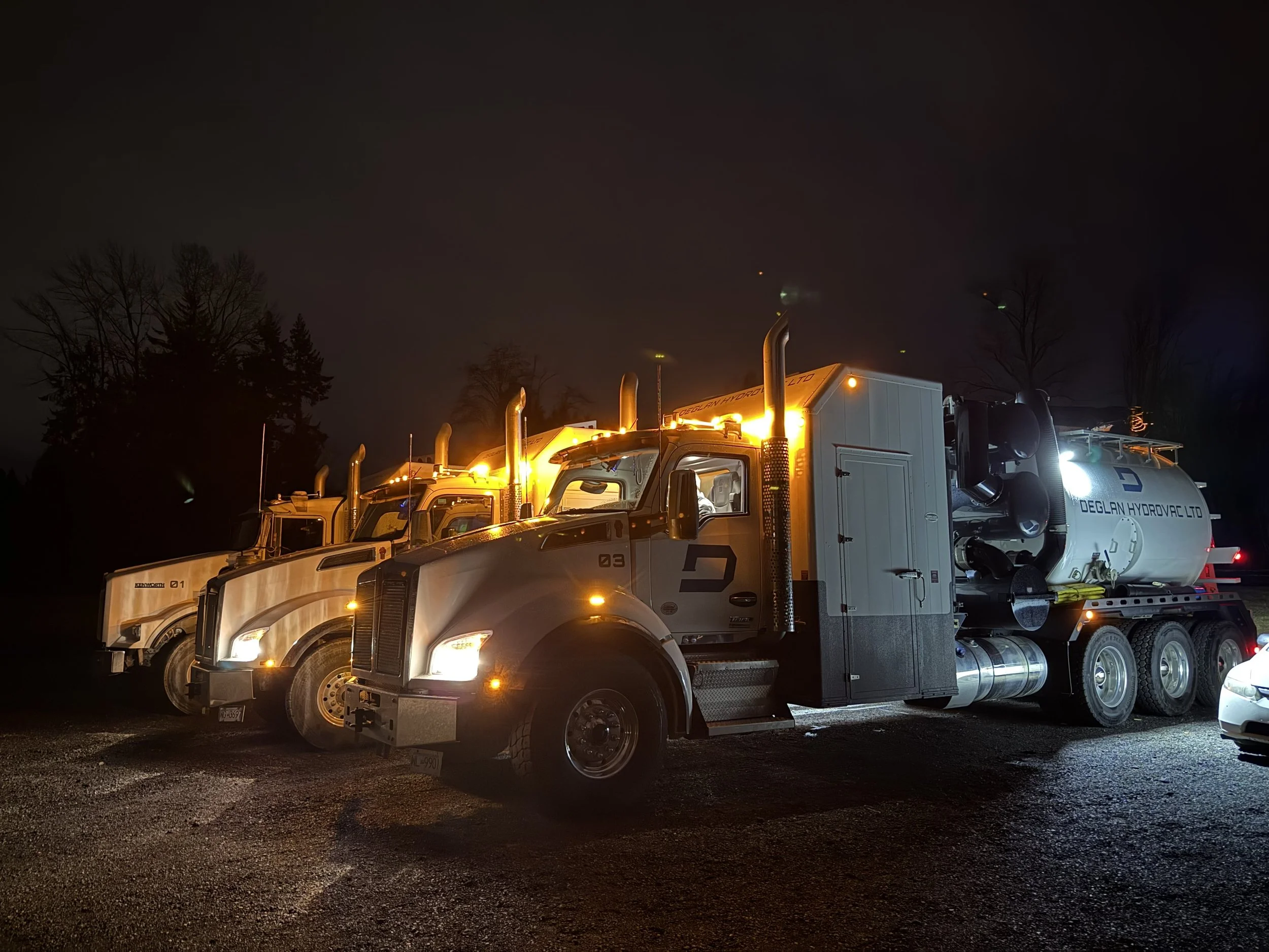 A line of three large white fire trucks with bright orange flashing lights parked on a dark night, with trees in the background.