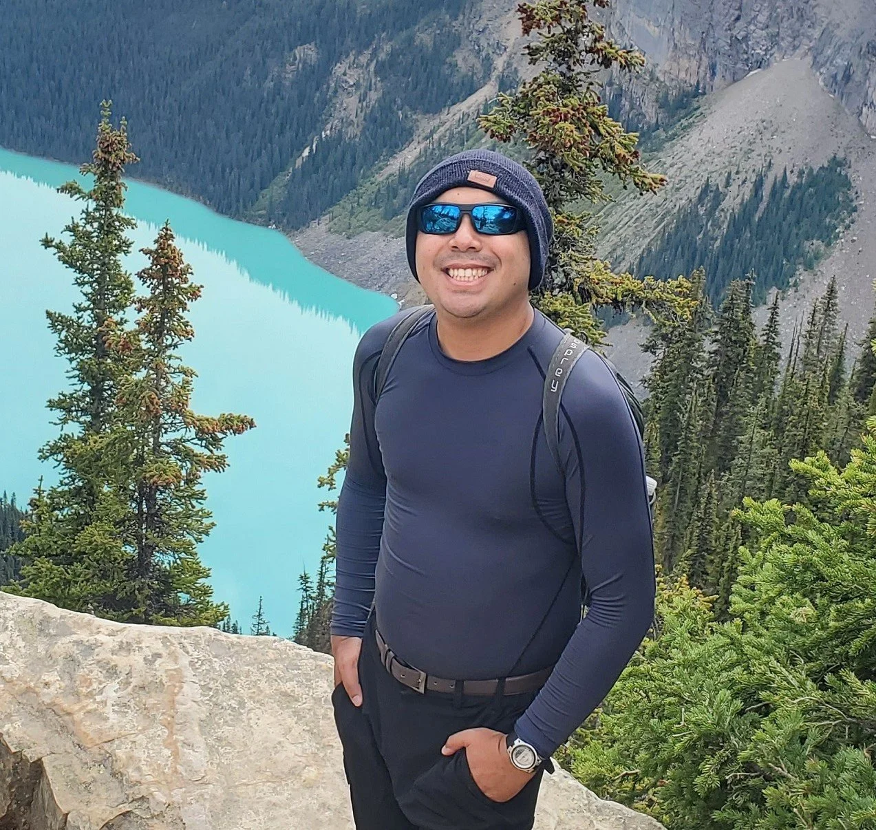 A man in outdoor hiking gear standing in front of a turquoise lake surrounded by green pine trees and mountains.