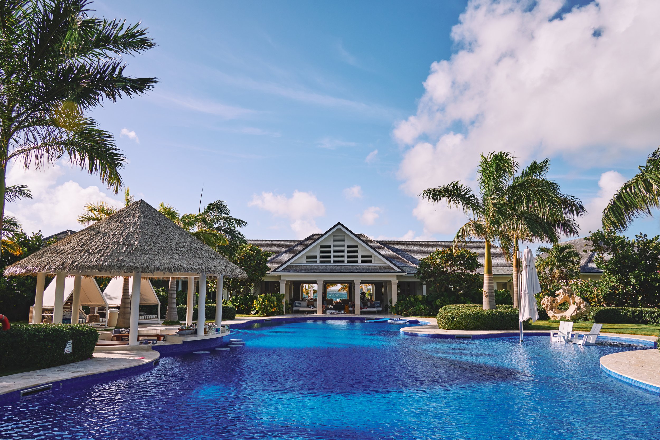Luxury resort swimming pool with thatched-roof cabana, palm trees, lounge chairs, and a building in the background under a partly cloudy sky.