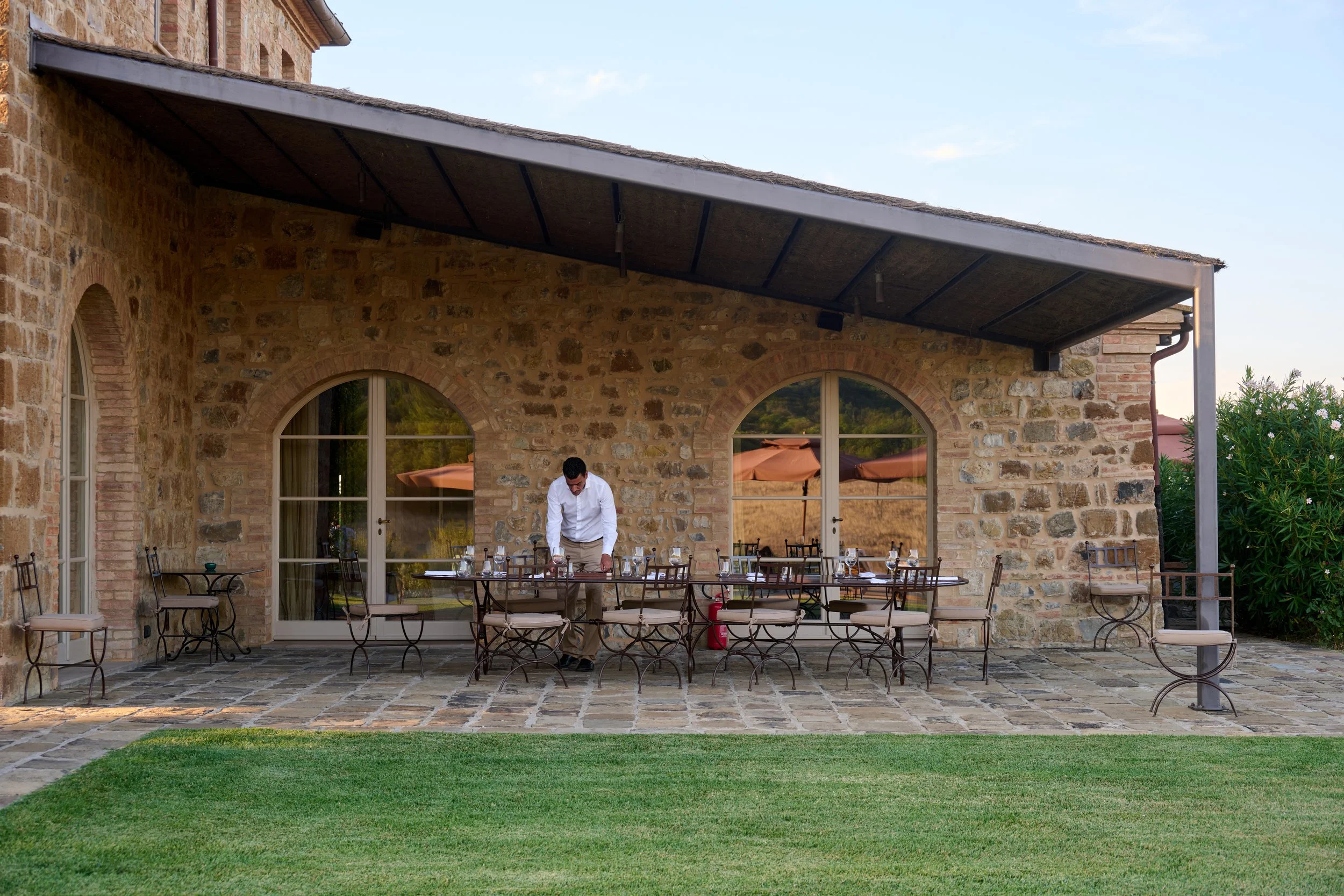 Man setting up an outdoor dining table on a stone patio in front of a stone building with large arched windows, under a roofed patio area.