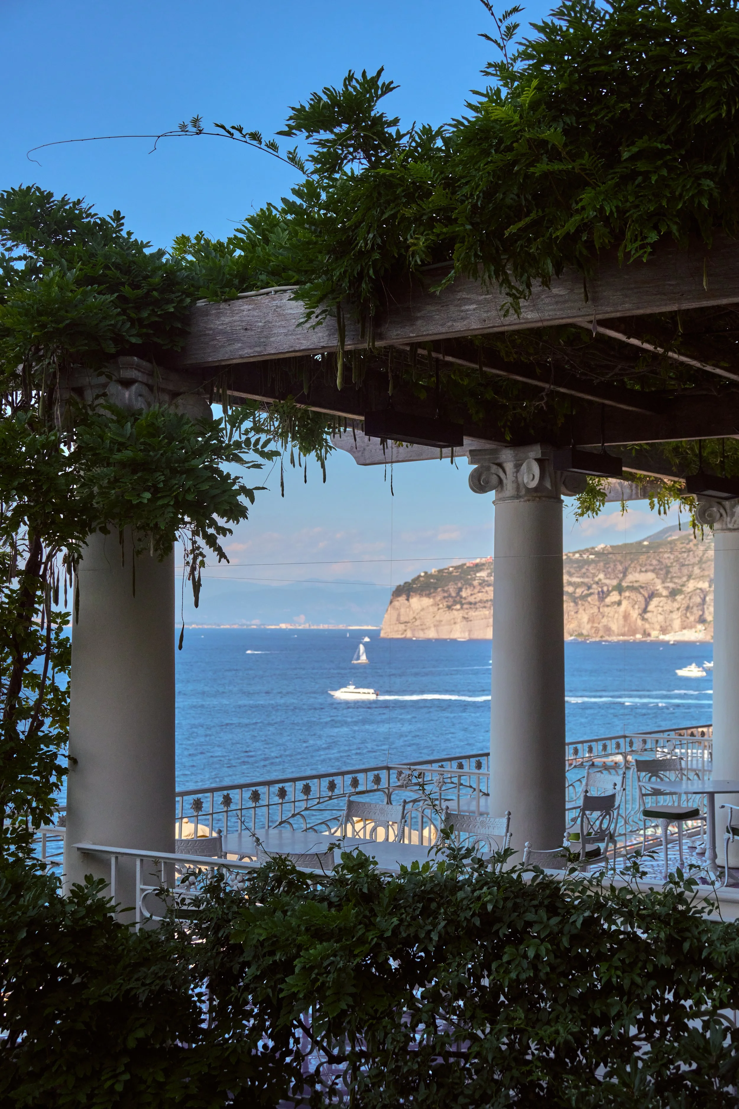 A view of a terrace overlooking the ocean with boats, framed by white columns and green foliage, under a wooden pergola.