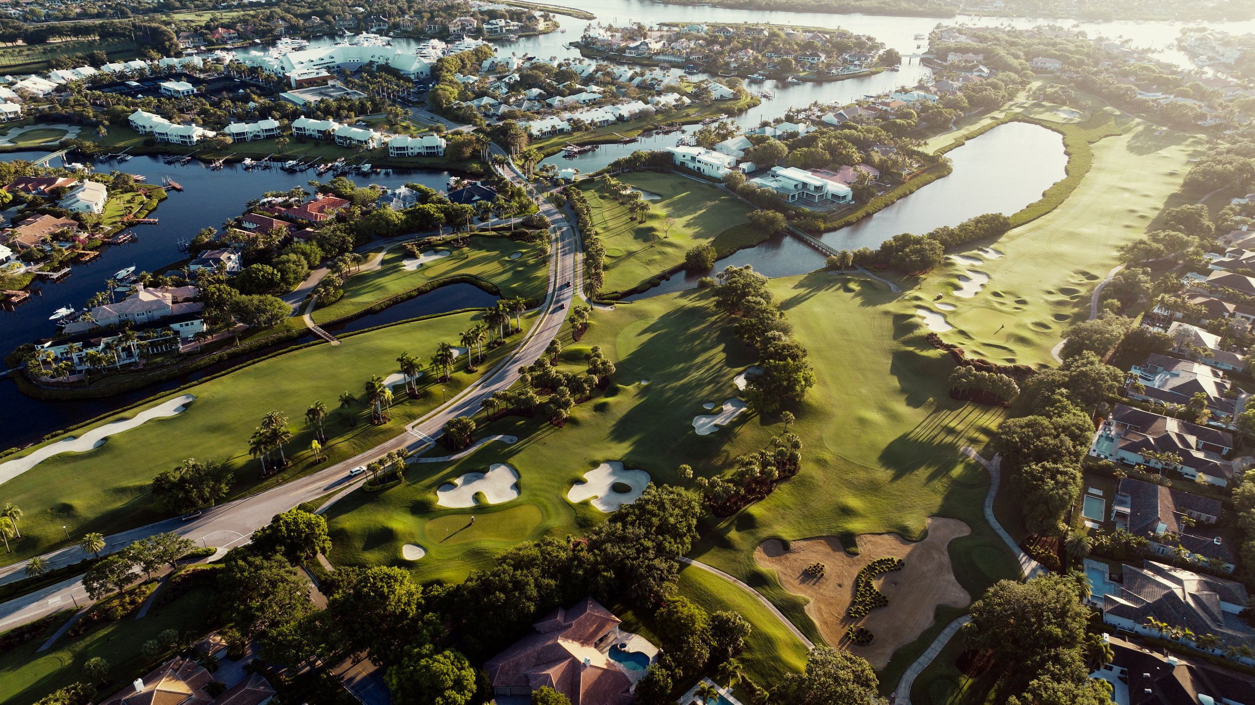 Aerial view of a golf course with sand traps, lakes, trees, and residential neighborhoods in the background.