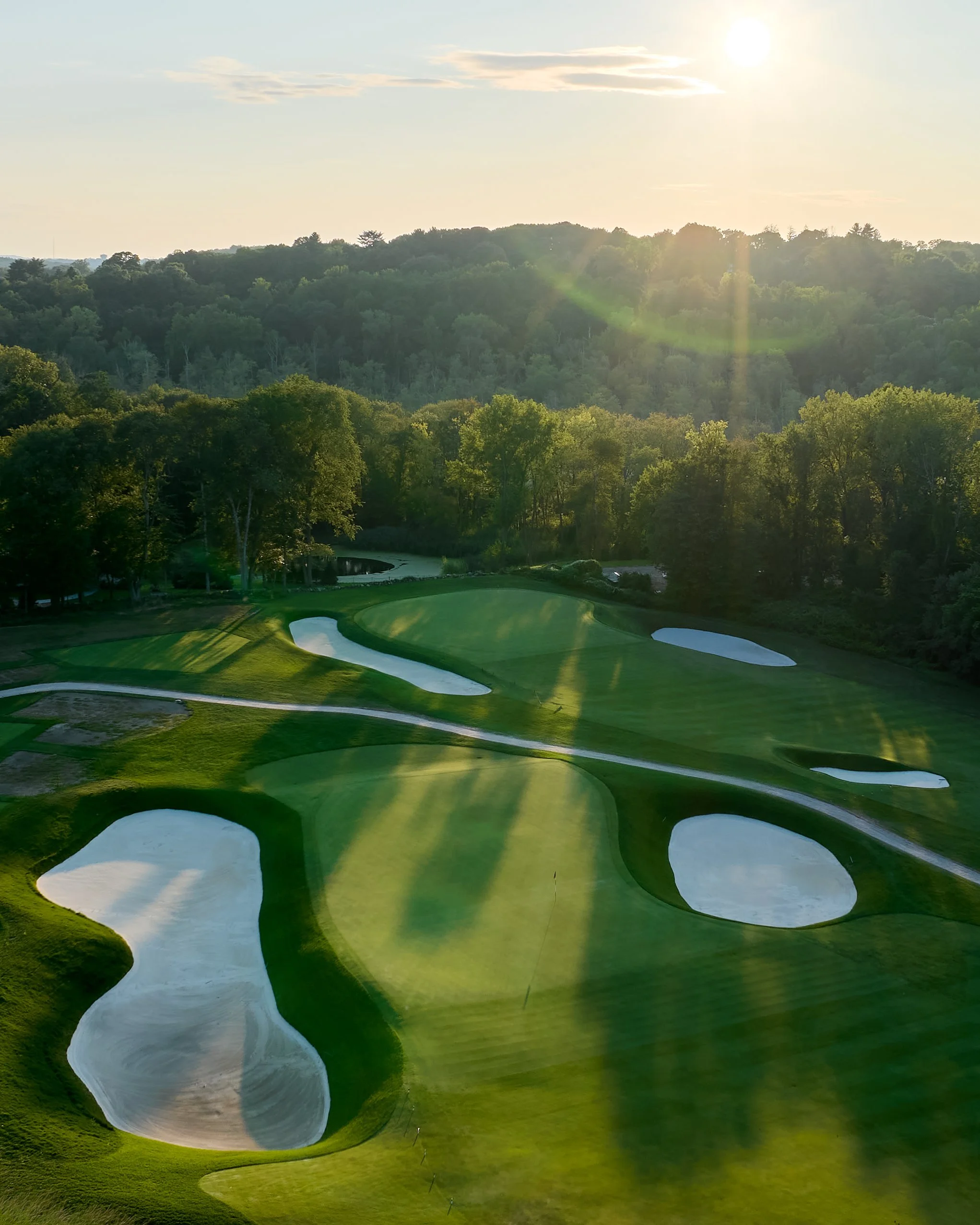 A golf course at sunset with sand traps and lush green fairways, surrounded by trees and rolling hills.