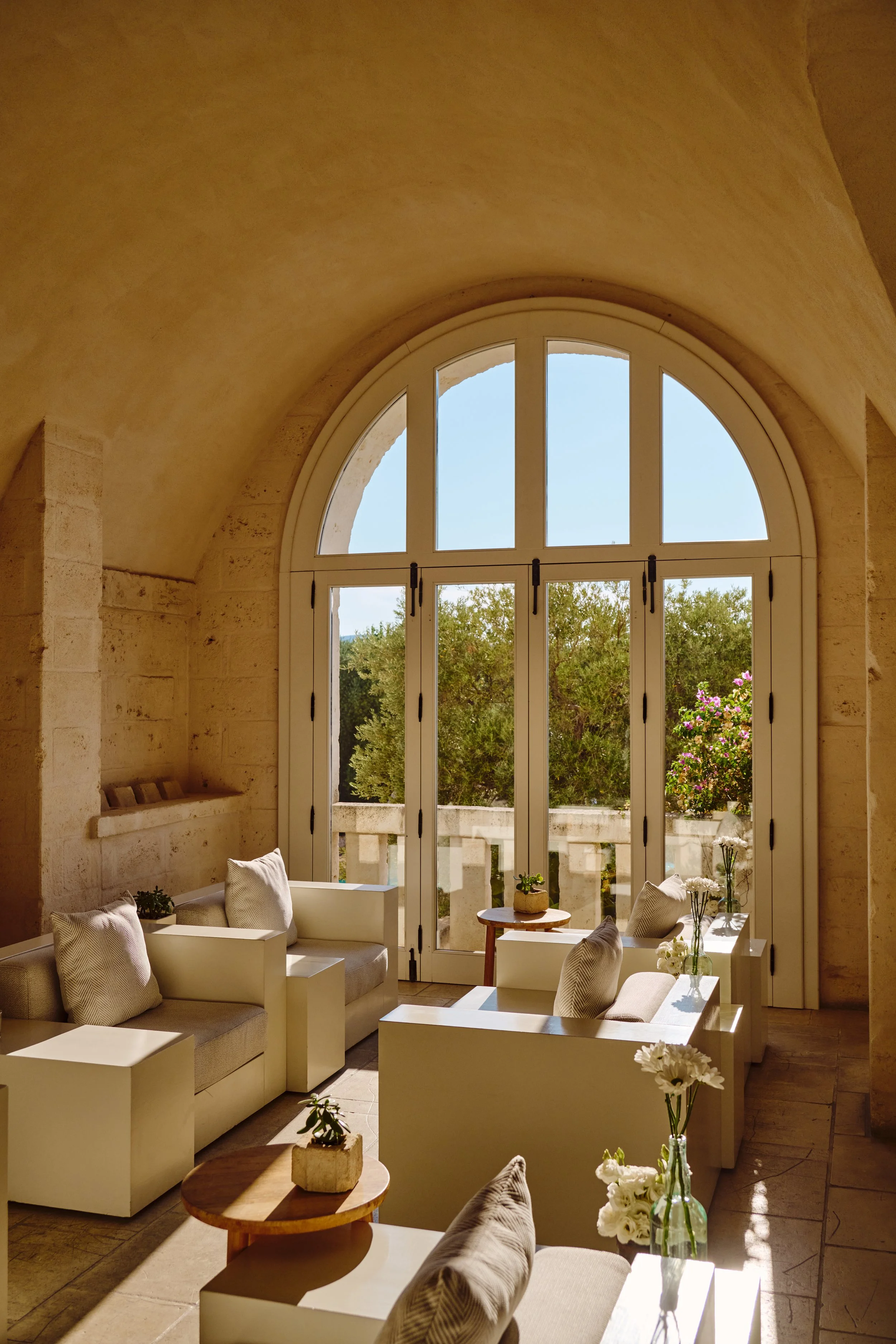 Sunlit indoor seating area with beige chairs, small wooden tables, and decorative plants, next to large arched windows showing trees and a balcony outside.