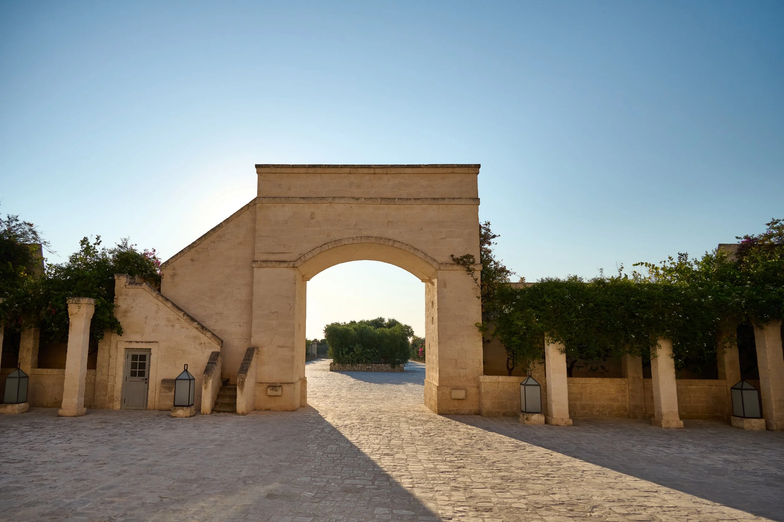 Stone archway and entrance with cobblestone pavement, flanked by greenery and lanterns, under a clear blue sky.
