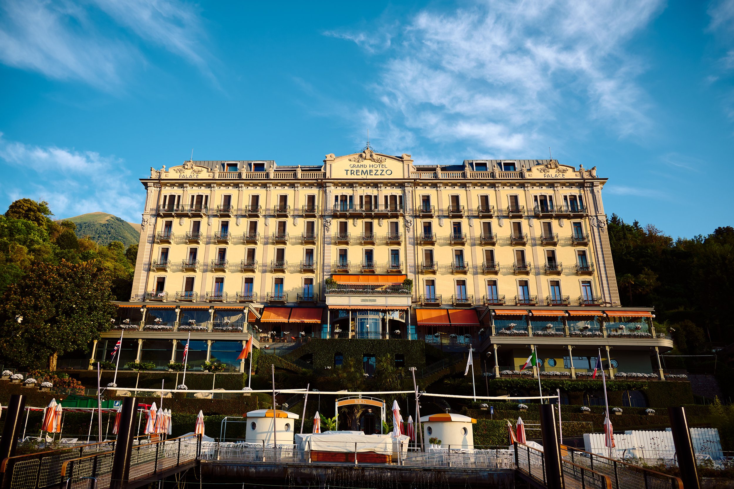 A large, multi-story hotel called Grand Hotel Tremezzo with a classic architecture, situated on a hillside with trees and mountains behind it, under a partly cloudy blue sky.