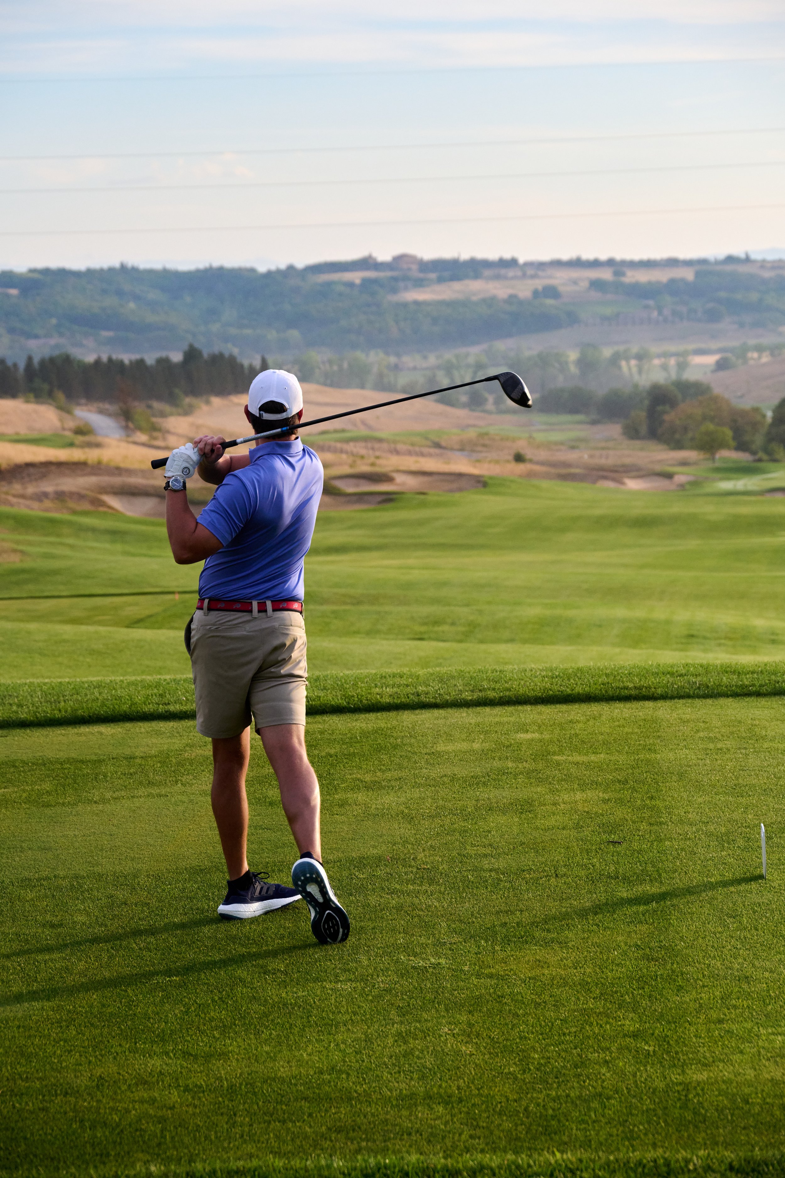 A man playing golf on a golf course, preparing to swing his club, with rolling hills and trees in the background.