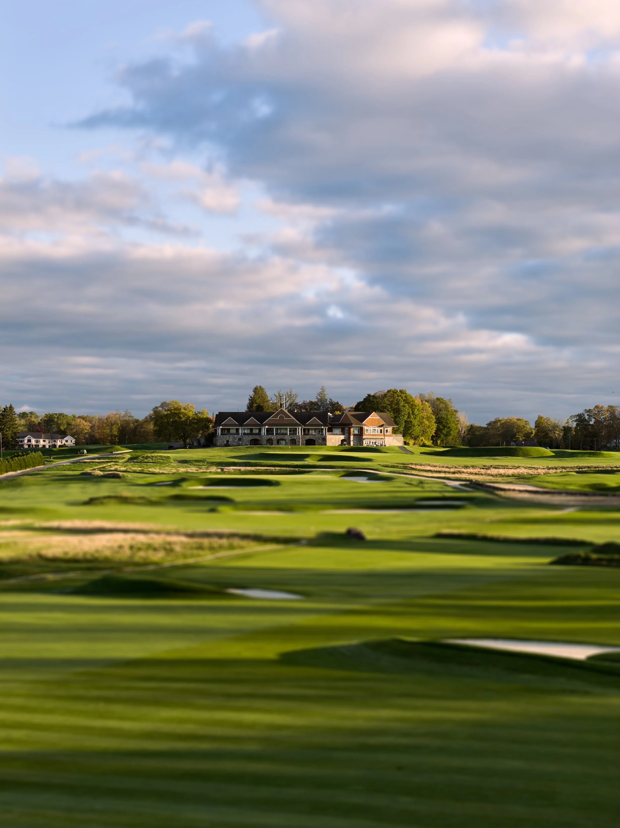 A golf course with green fairways, sand traps, and a large house in the background under a partly cloudy sky.