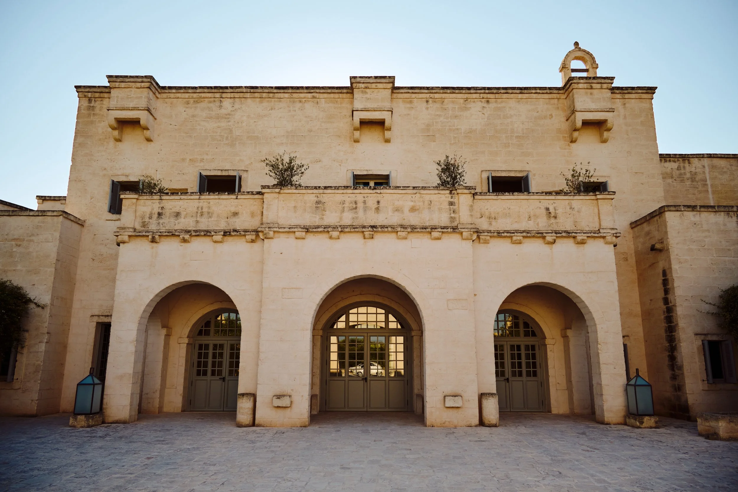 A historic stone building with three arched entrances, two lanterns on either side, and small trees planted on the second-floor balcony, under a clear blue sky.