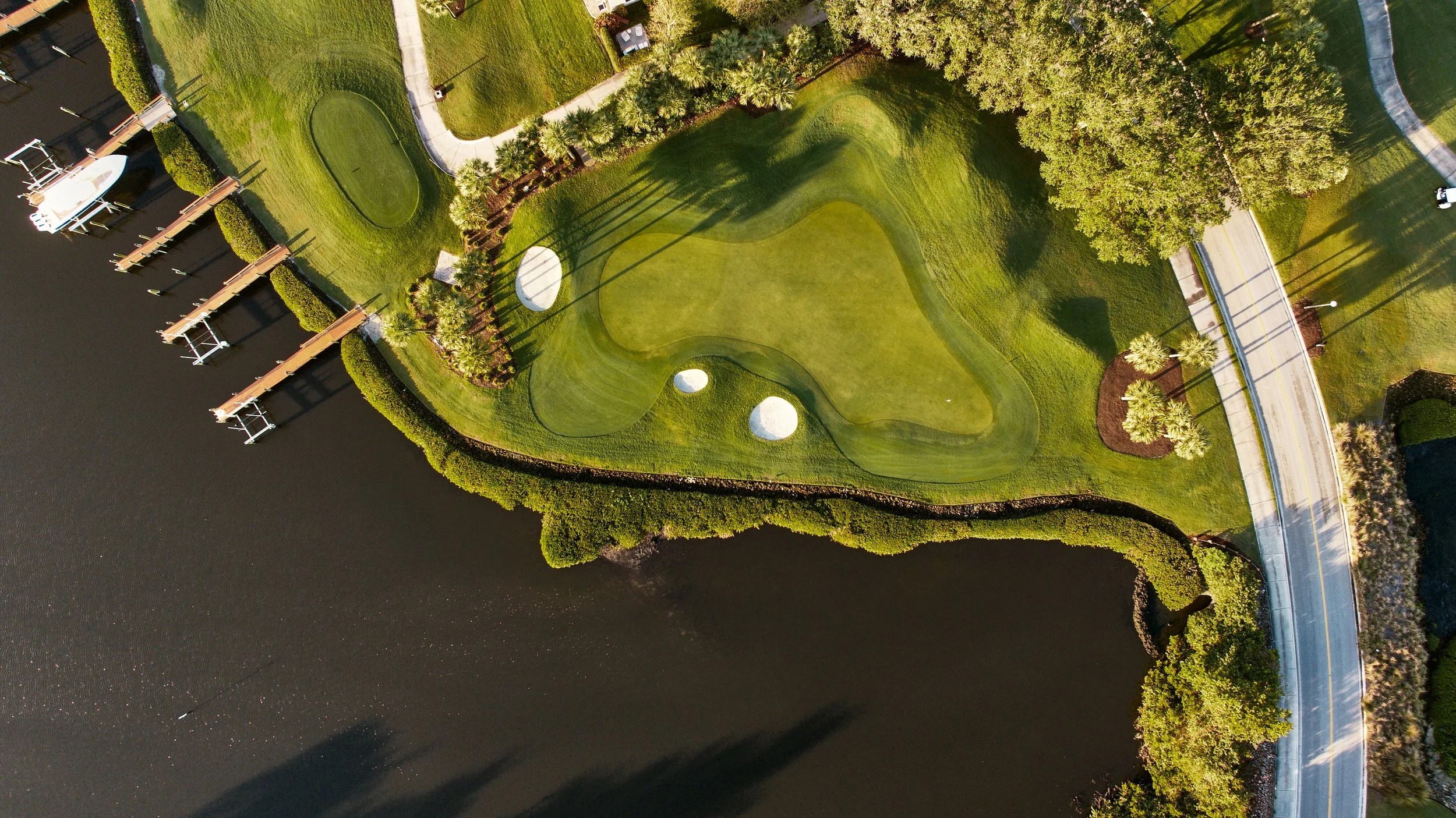 An aerial view of a golf course with a putting green, sand bunkers, trees, a road, and a body of water with a dock.