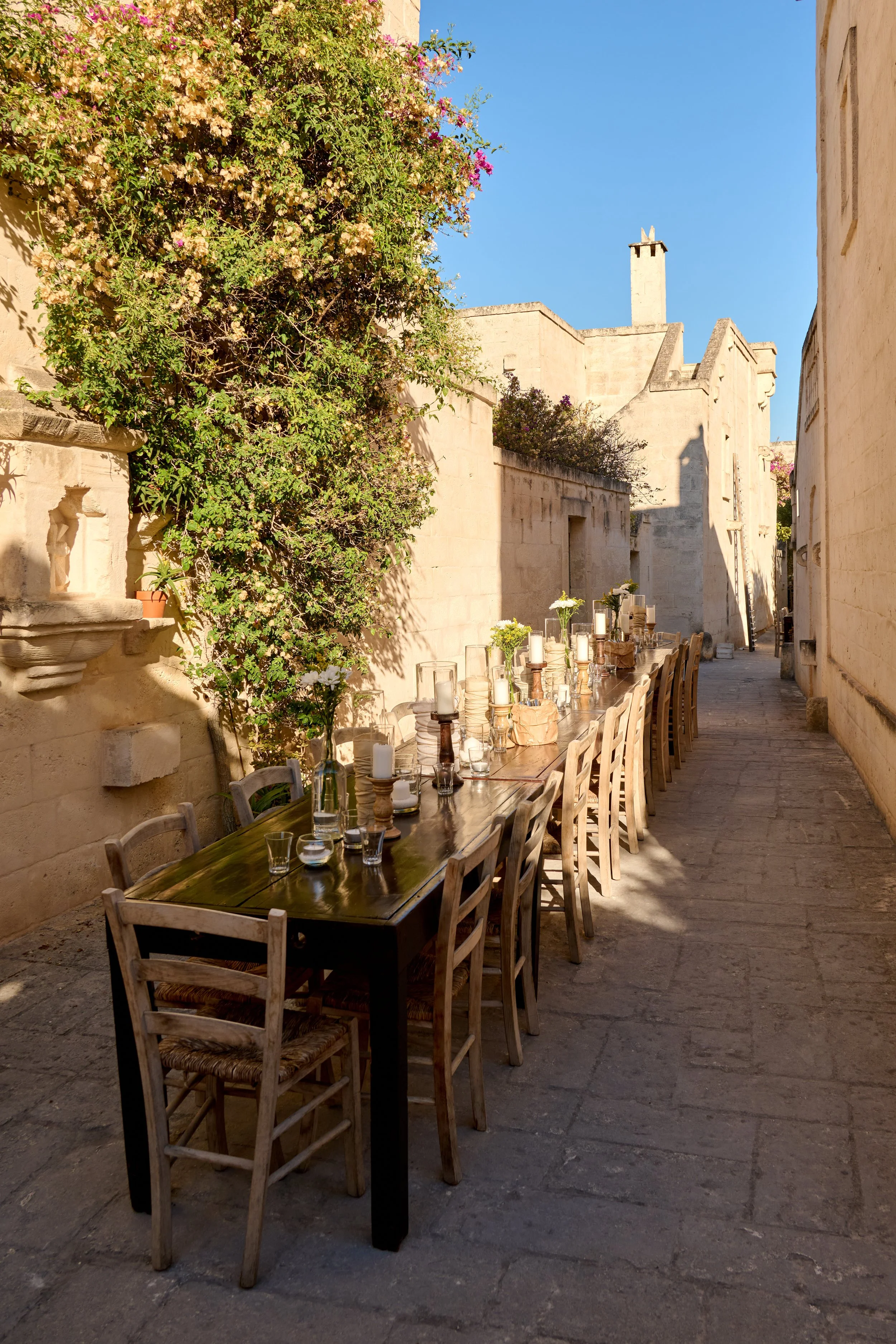 Outdoor dining table set along a narrow stone alleyway with chairs, candles, and flower vases, surrounded by stone buildings and greenery, under a clear blue sky.