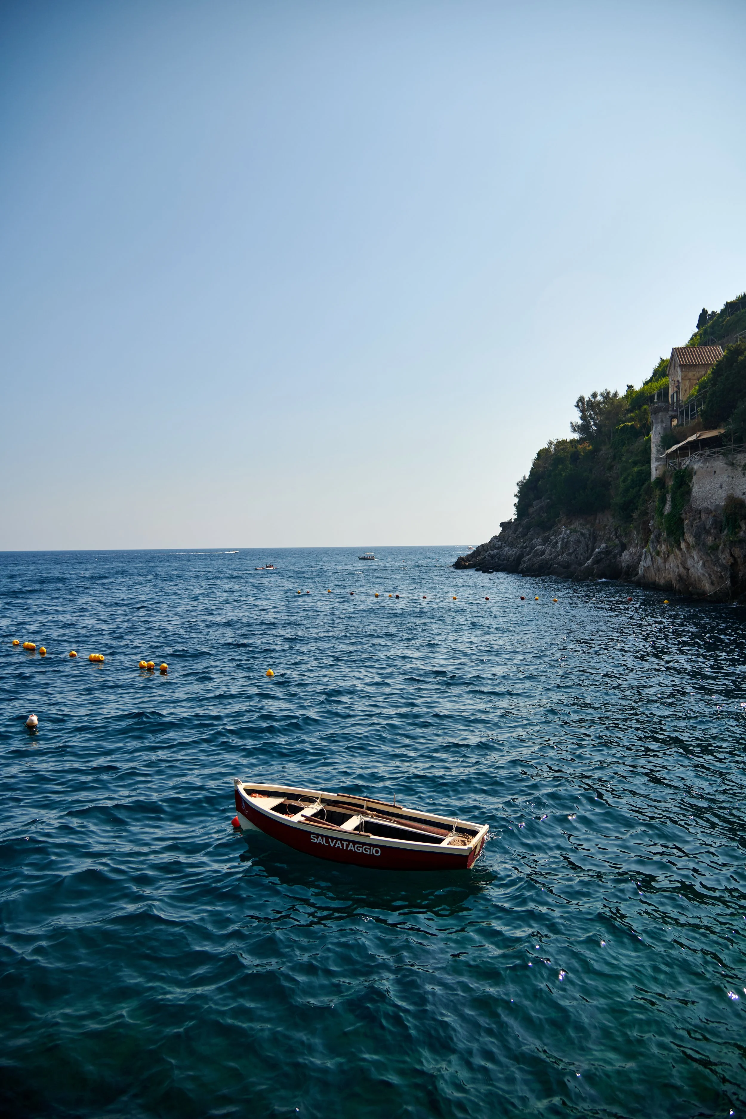 A small boat named Salvataggio floating on the ocean near a rocky coastline with houses and lush greenery.