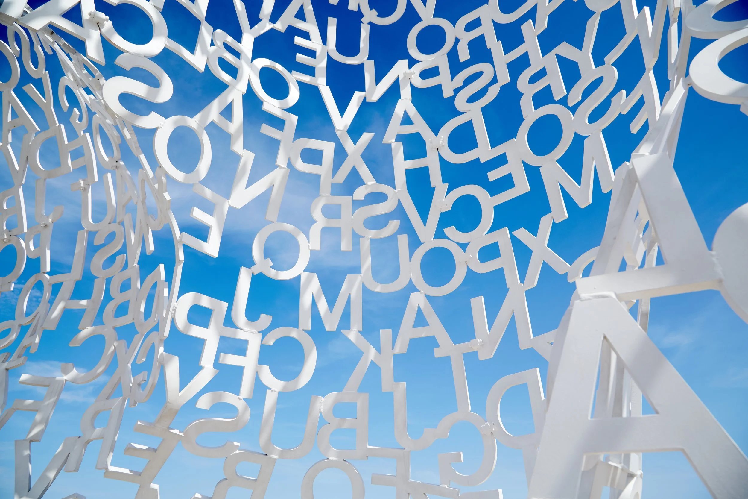 A modern outdoor sculpture made of white interconnected alphabetic letters against a blue sky with clouds.