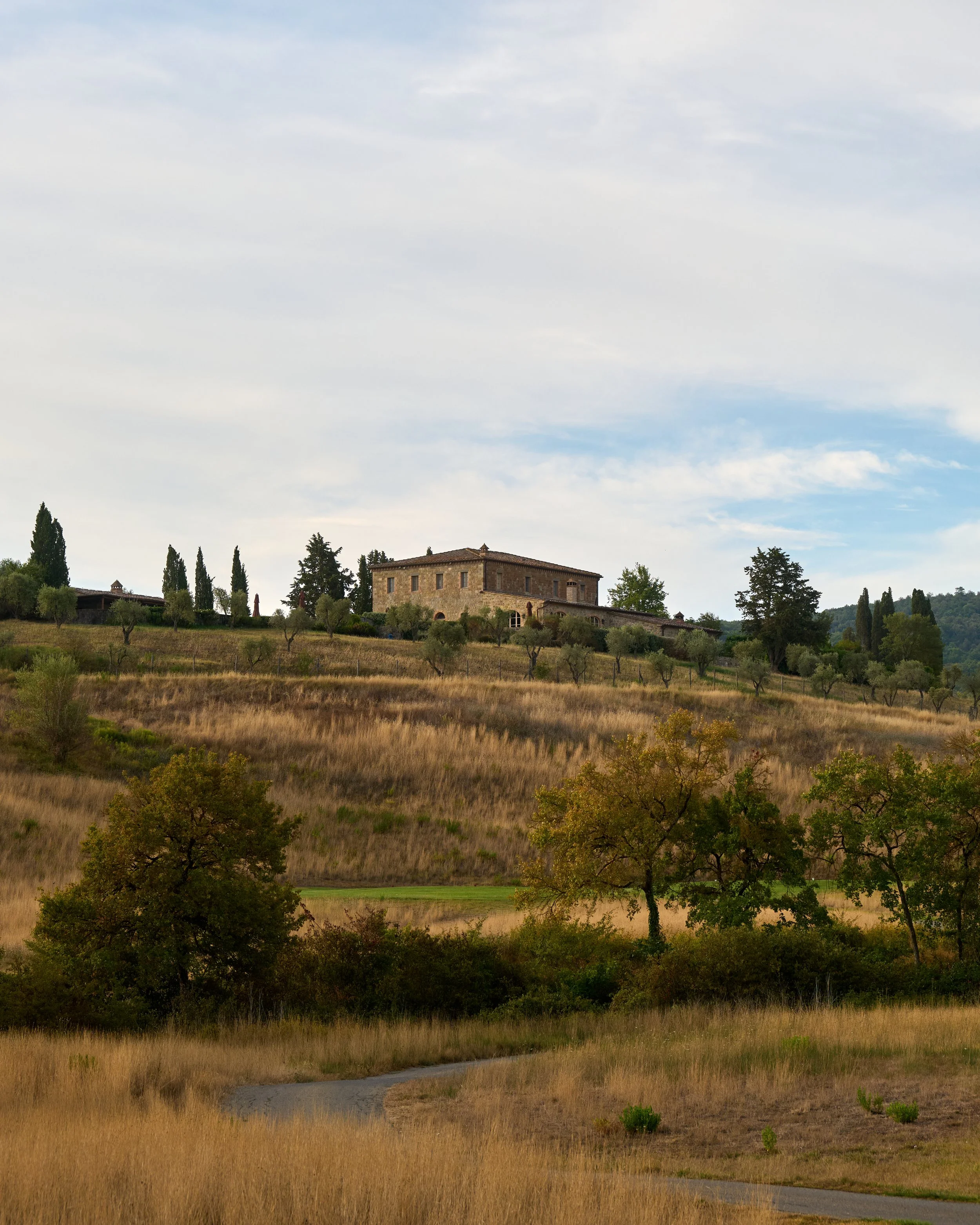 Scenic view of a hilly countryside with a stone house on top, surrounded by trees and open fields, under a partly cloudy sky.