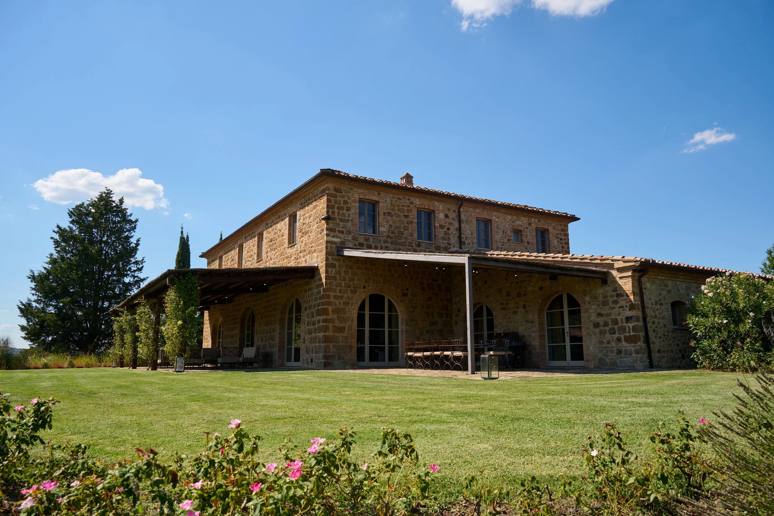 A large stone house with a tiled roof, surrounded by a well-maintained lawn and some flowering bushes, under a blue sky with a few clouds.