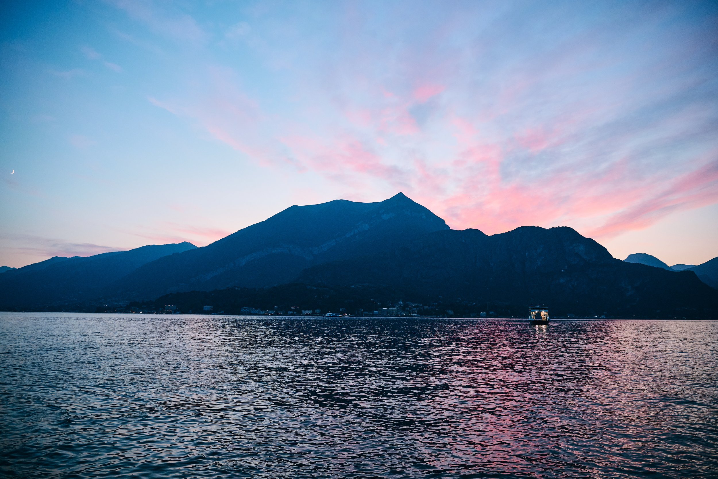 A scenic view of a mountain landscape during sunset, with a large body of water in the foreground and a boat sailing on the lake.