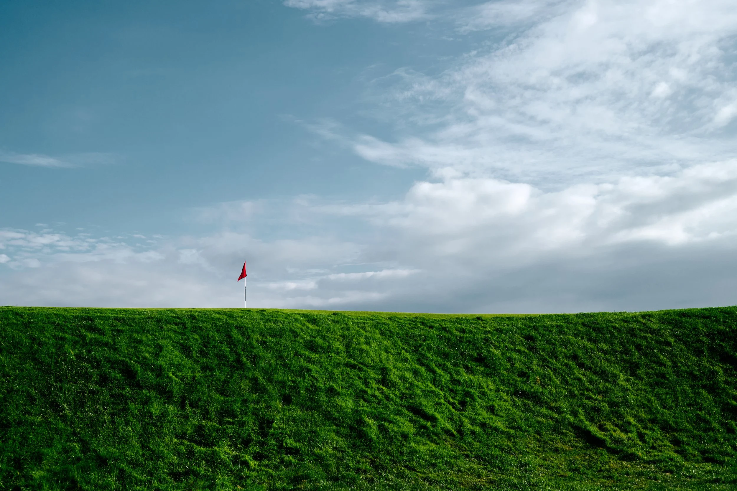 A green grassy hill with a red flag at the top, under a partly cloudy blue sky.