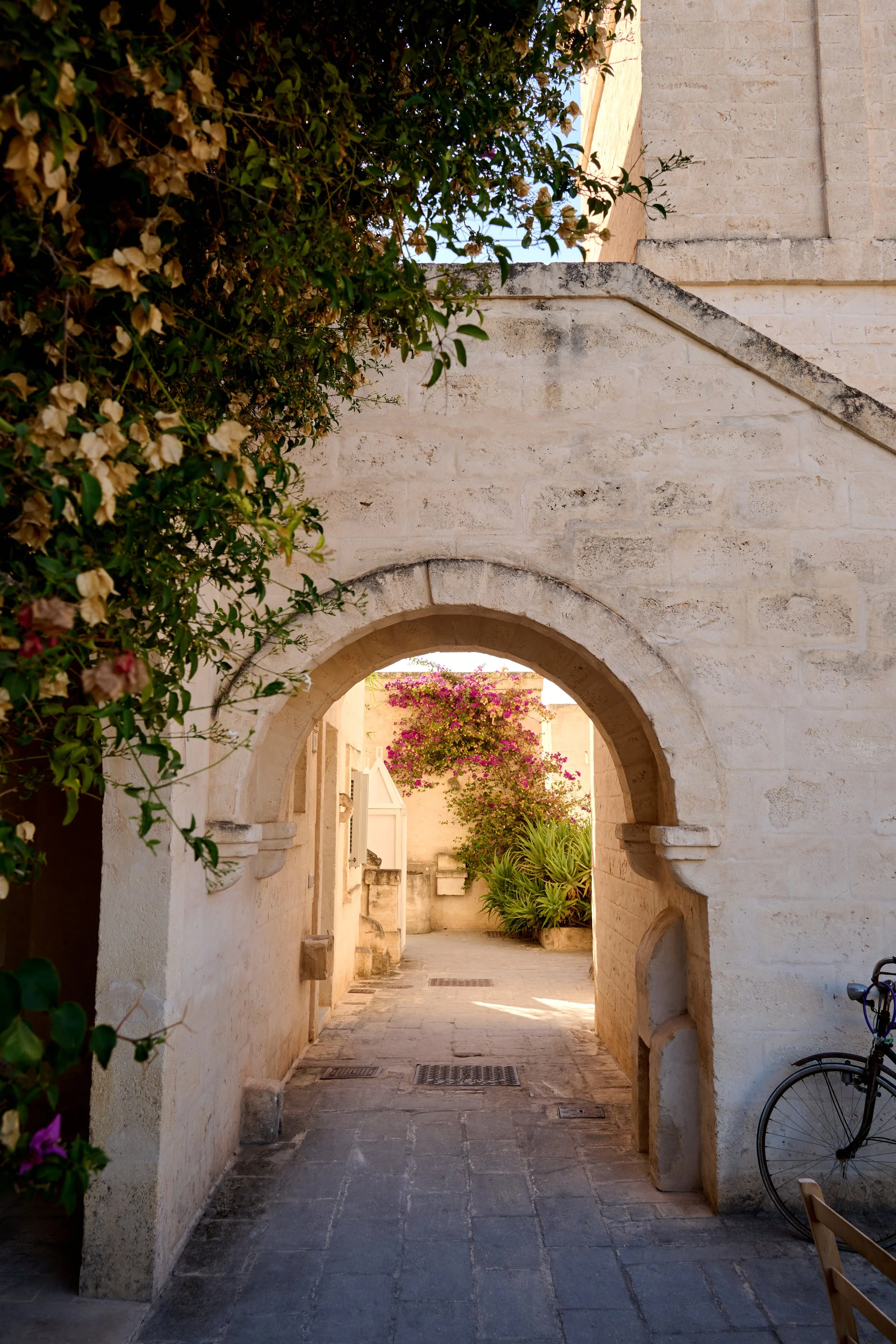 A narrow stone alleyway framed by an arched stone doorway with blooming pink bougainvillaea and green plants in the background, with a bicycle on the right side.