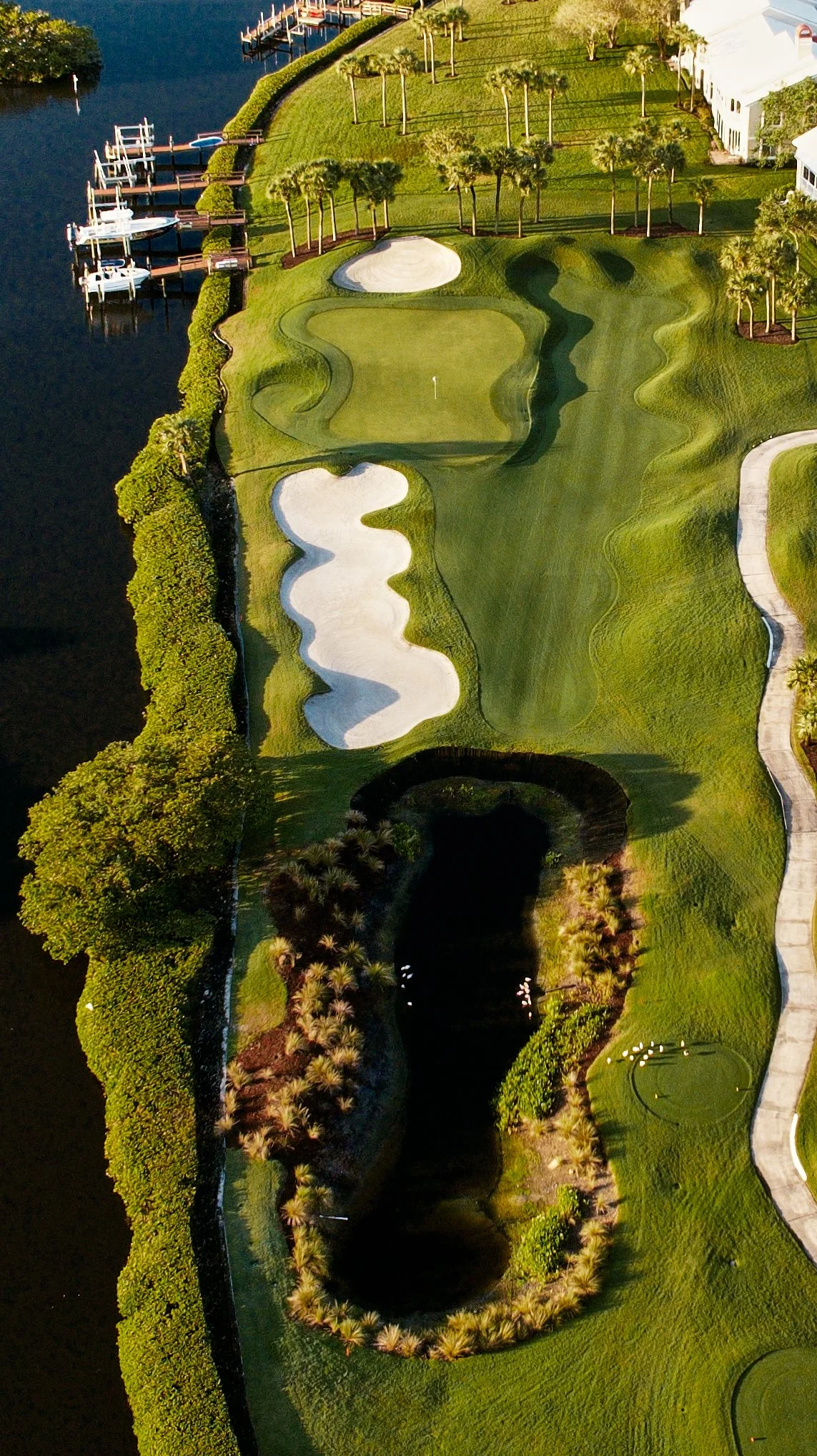 Aerial view of a golf course with a water hazard, sand bunkers, green fairways, and surrounding trees near a body of water with docks and boats.