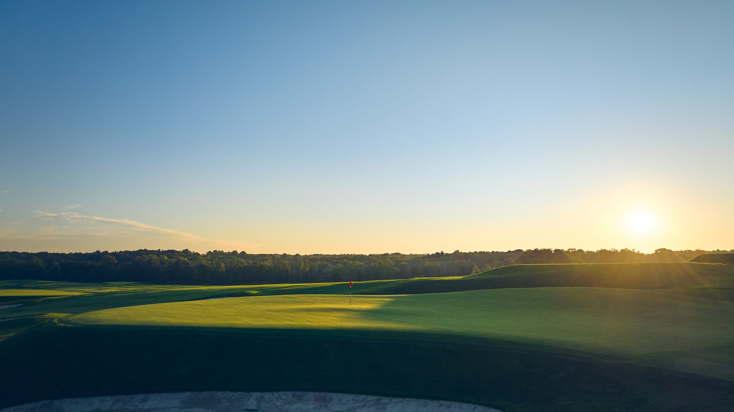 A golf course at sunset with a green, flag, trees in the background, and the sun setting on the horizon.