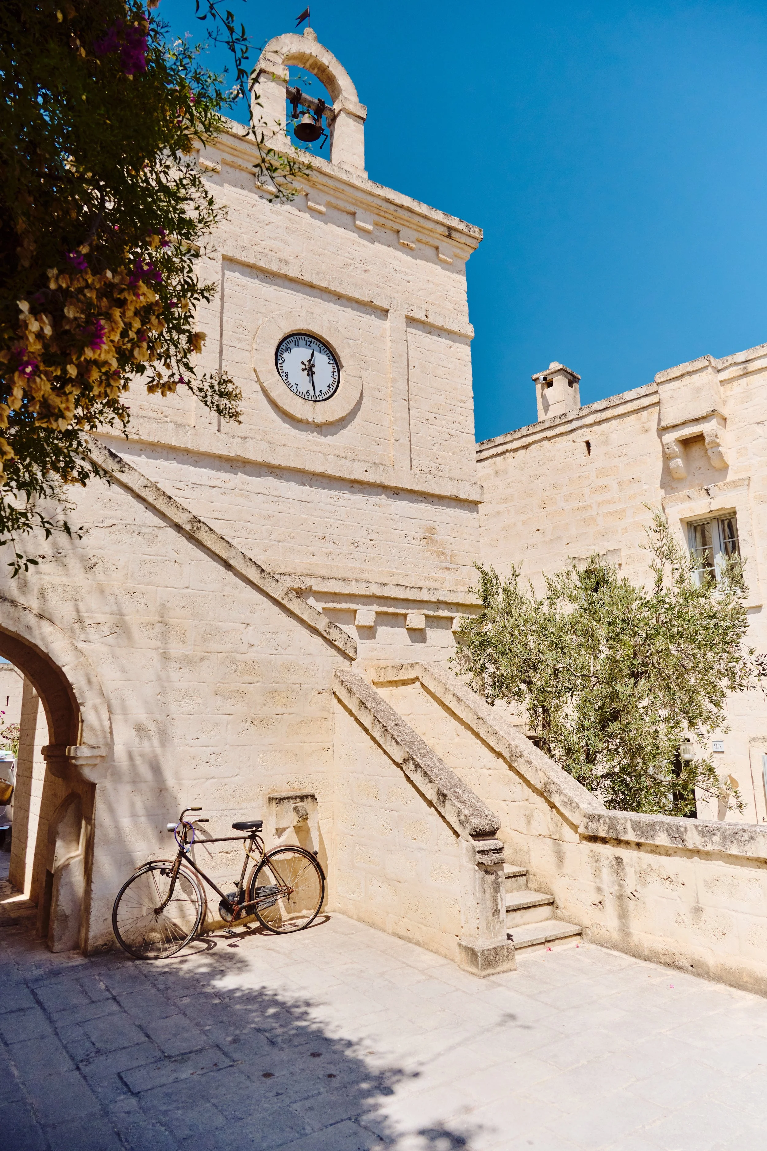 A historic stone building with a clock tower, a bike leaning against the wall, and a staircase leading up from the street in a sunny, clear sky setting.
