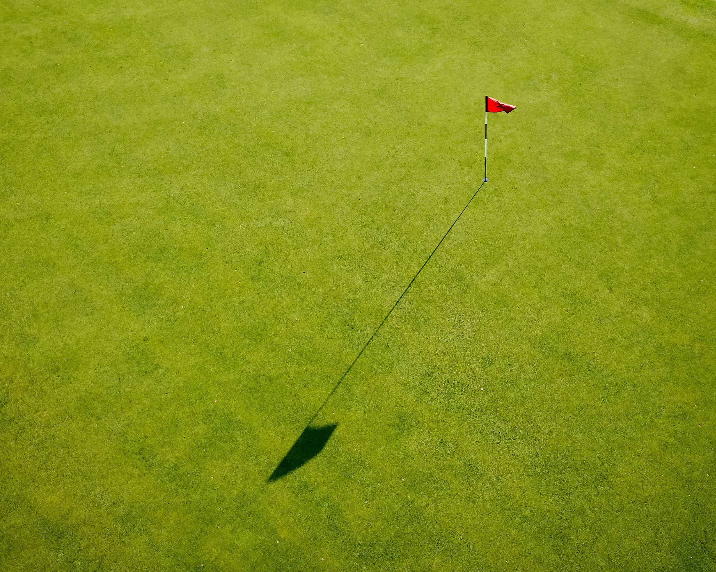 A golf green with a red flag marking the hole, casting a shadow.