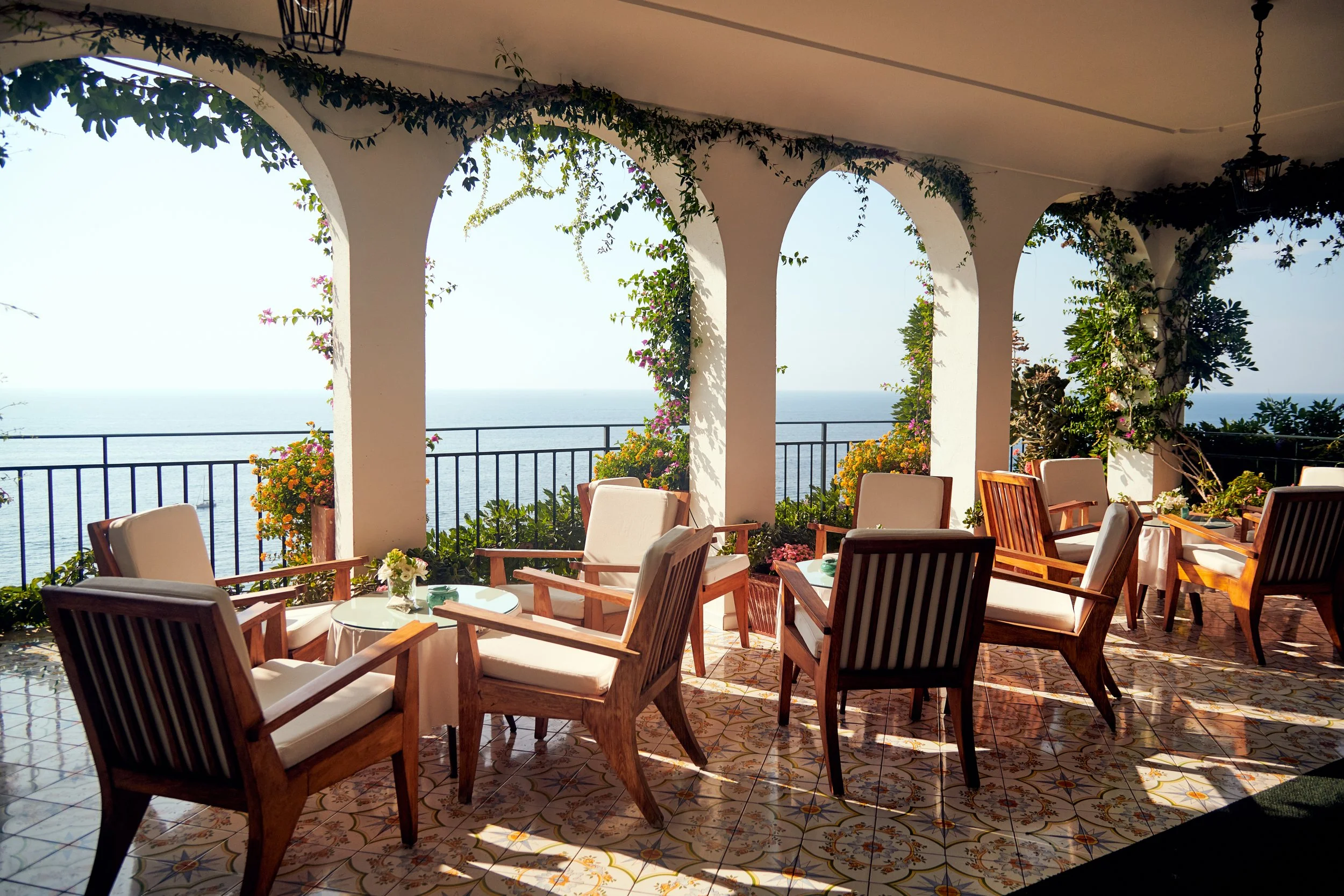 Outdoor terrace with wooden chairs and tables, overlooking the ocean with potted plants and flowering vines along the white arched columns.