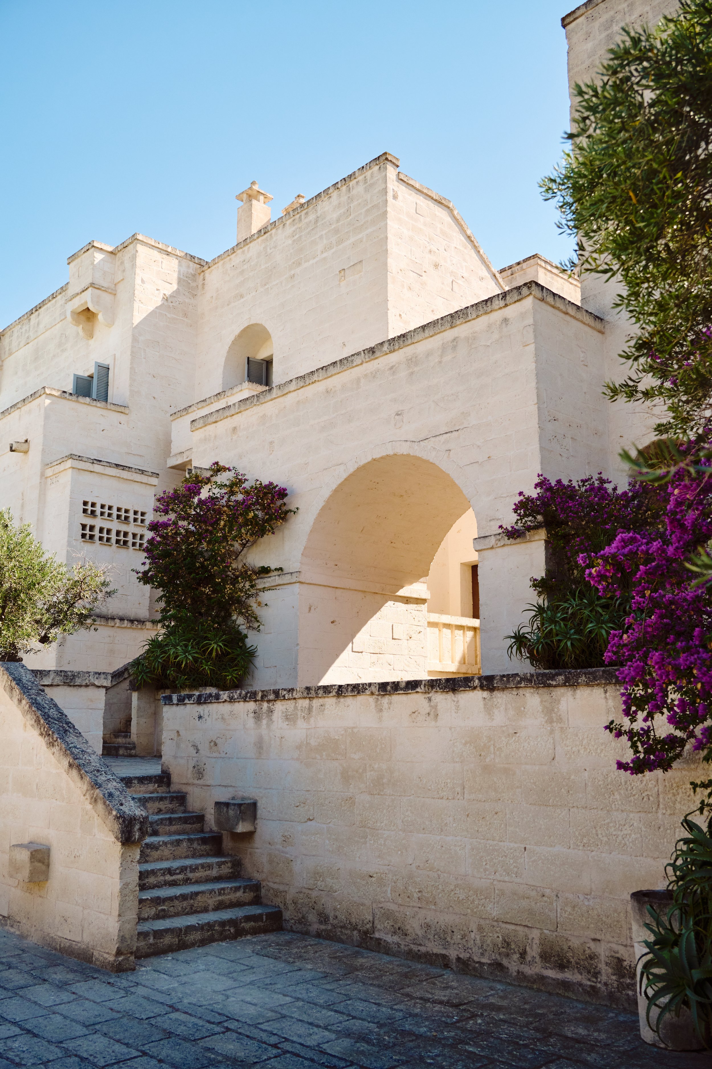 White stone building with an arched entrance, surrounded by purple flowers and greenery under a clear blue sky.