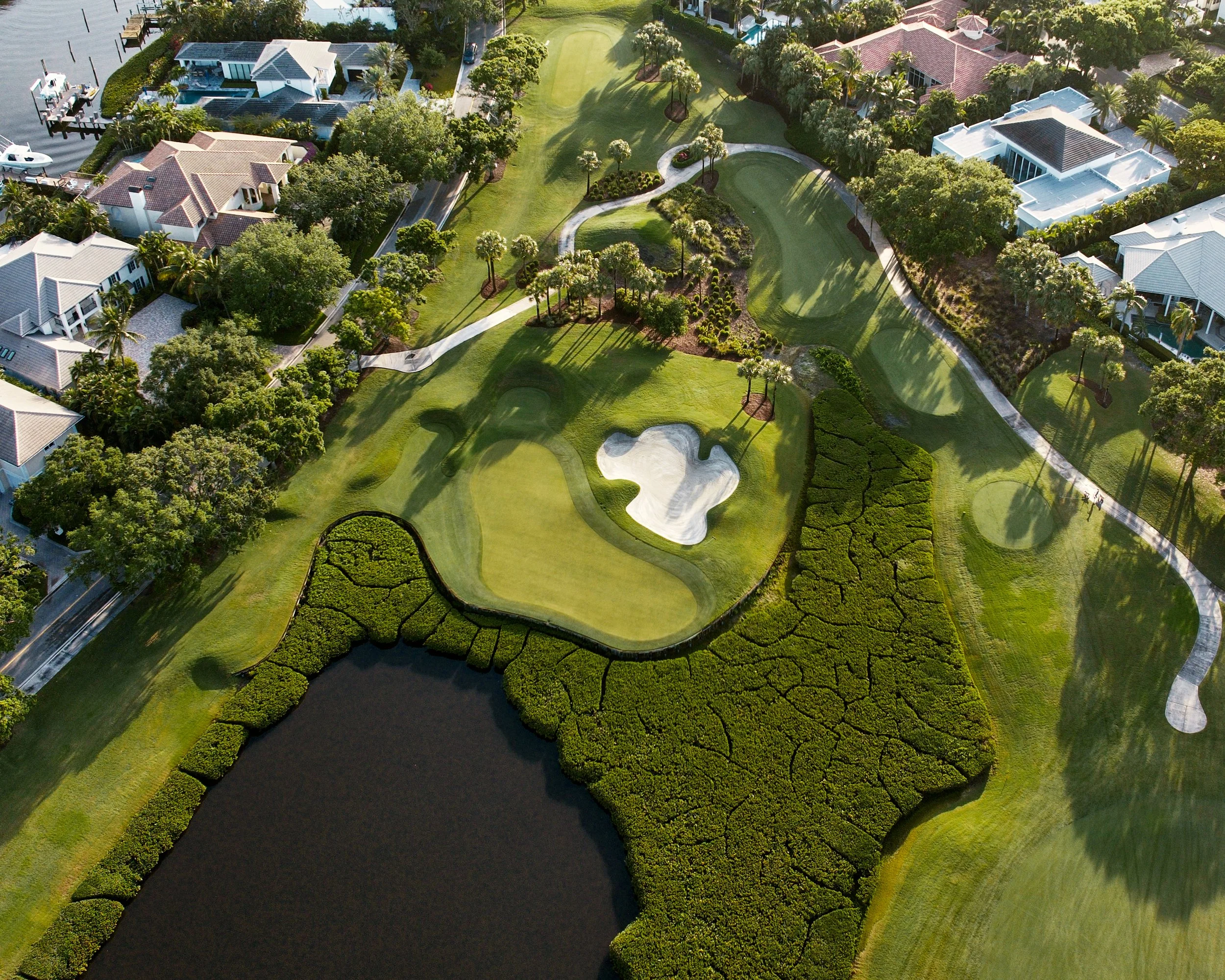 An aerial view of a golf course surrounded by residential homes, featuring green fairways, a sand bunker, a water hazard, and walking paths, with trees casting shadows across the landscape.