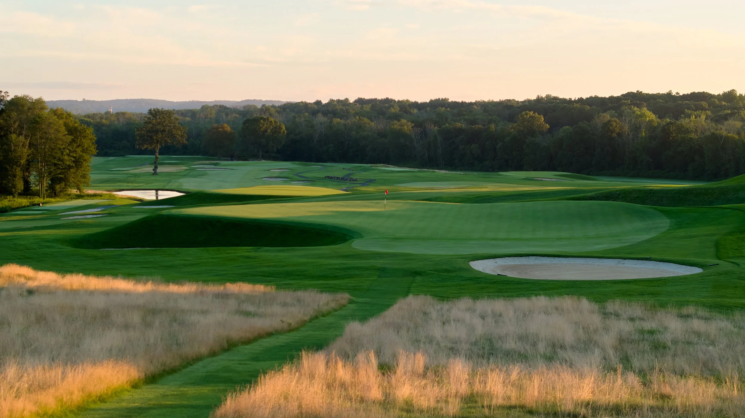 A scenic golf course with green fairways, sand bunkers, and a red flag on the putting green, surrounded by trees and open space during sunlight.