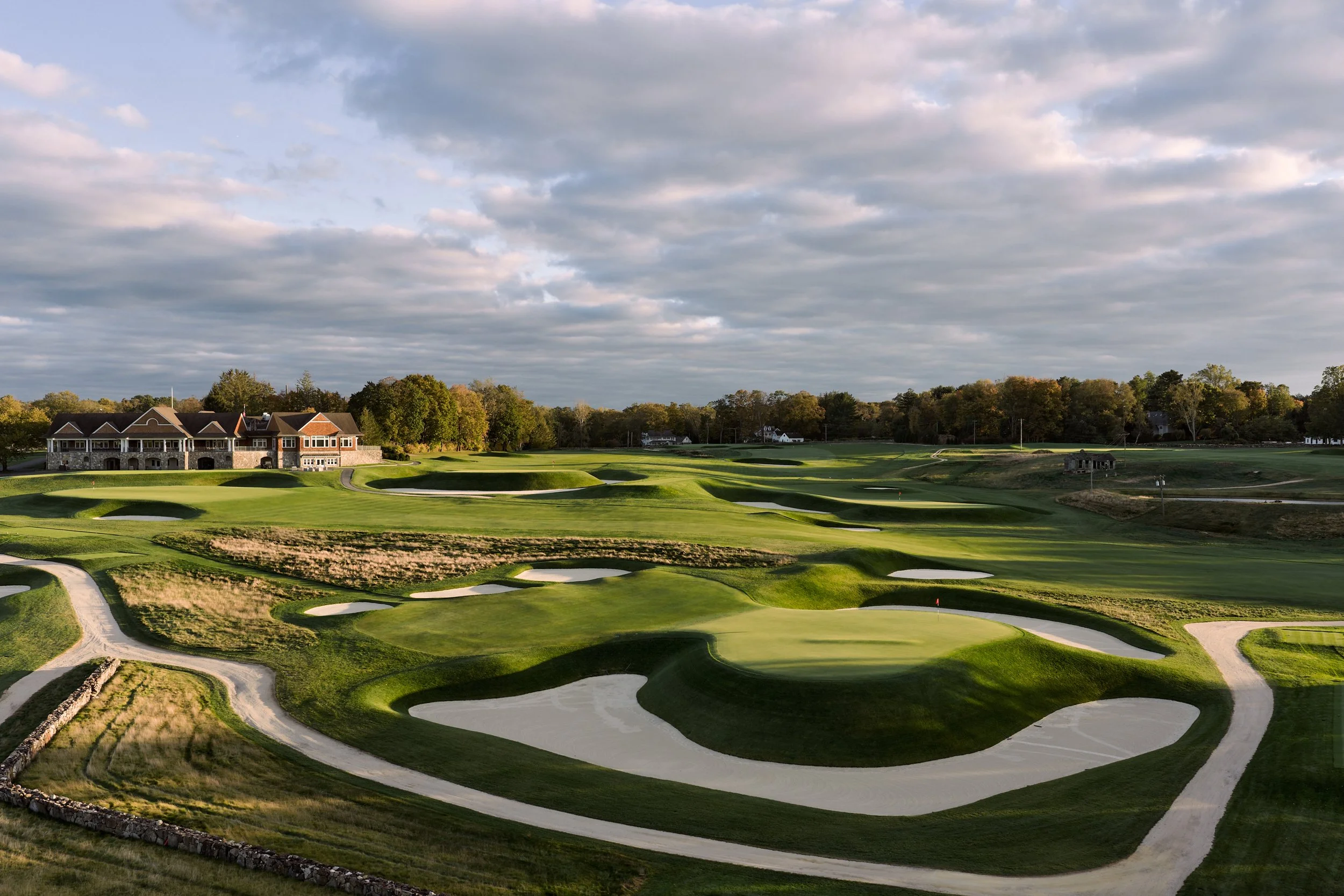 A golf course with sand traps, putting greens, and a large building in the background under a cloudy sky