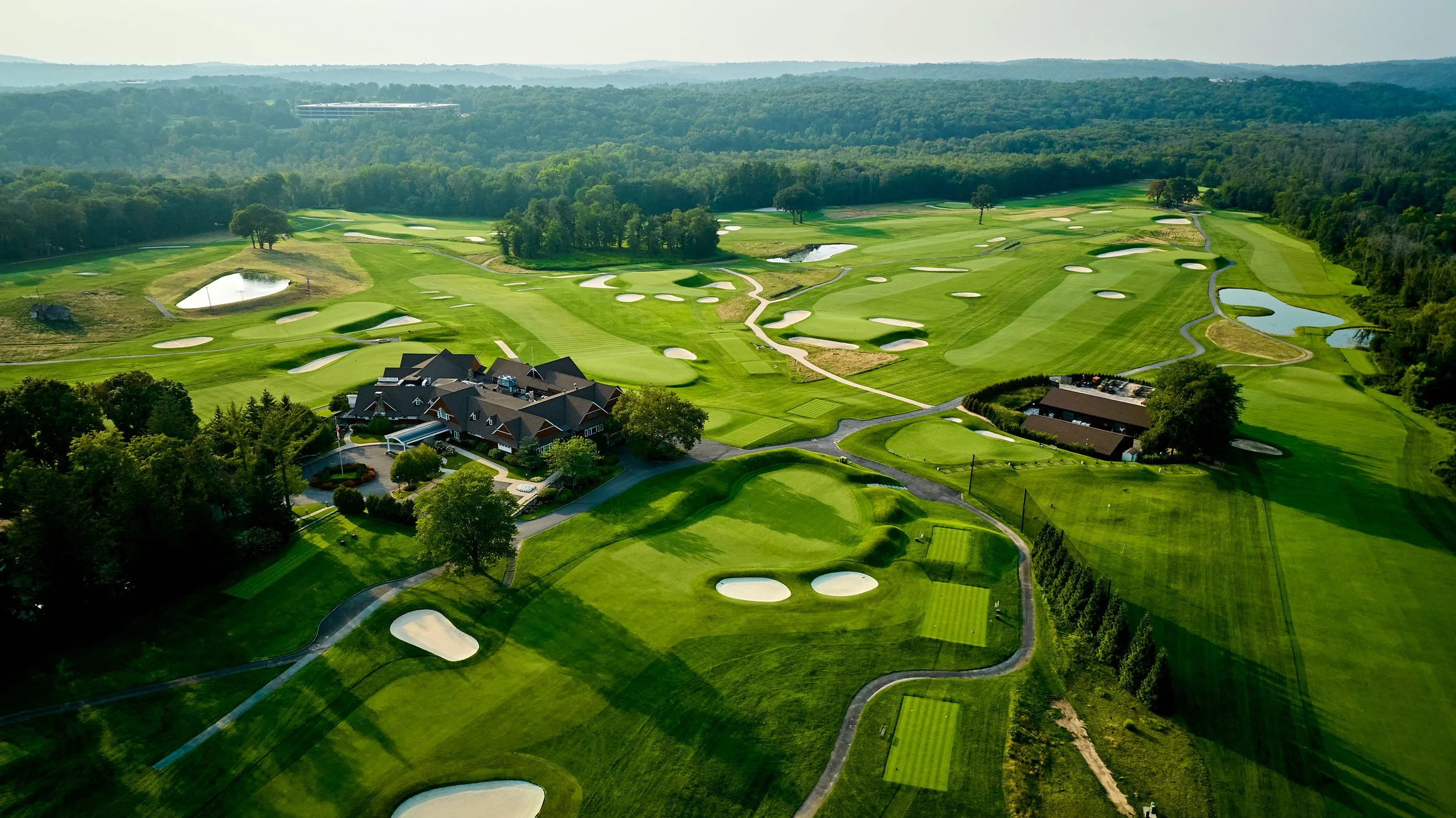 Aerial view of a golf course with multiple sand traps, water hazards, and well-maintained greens surrounded by trees and rolling landscape, with clubhouse buildings nearby.