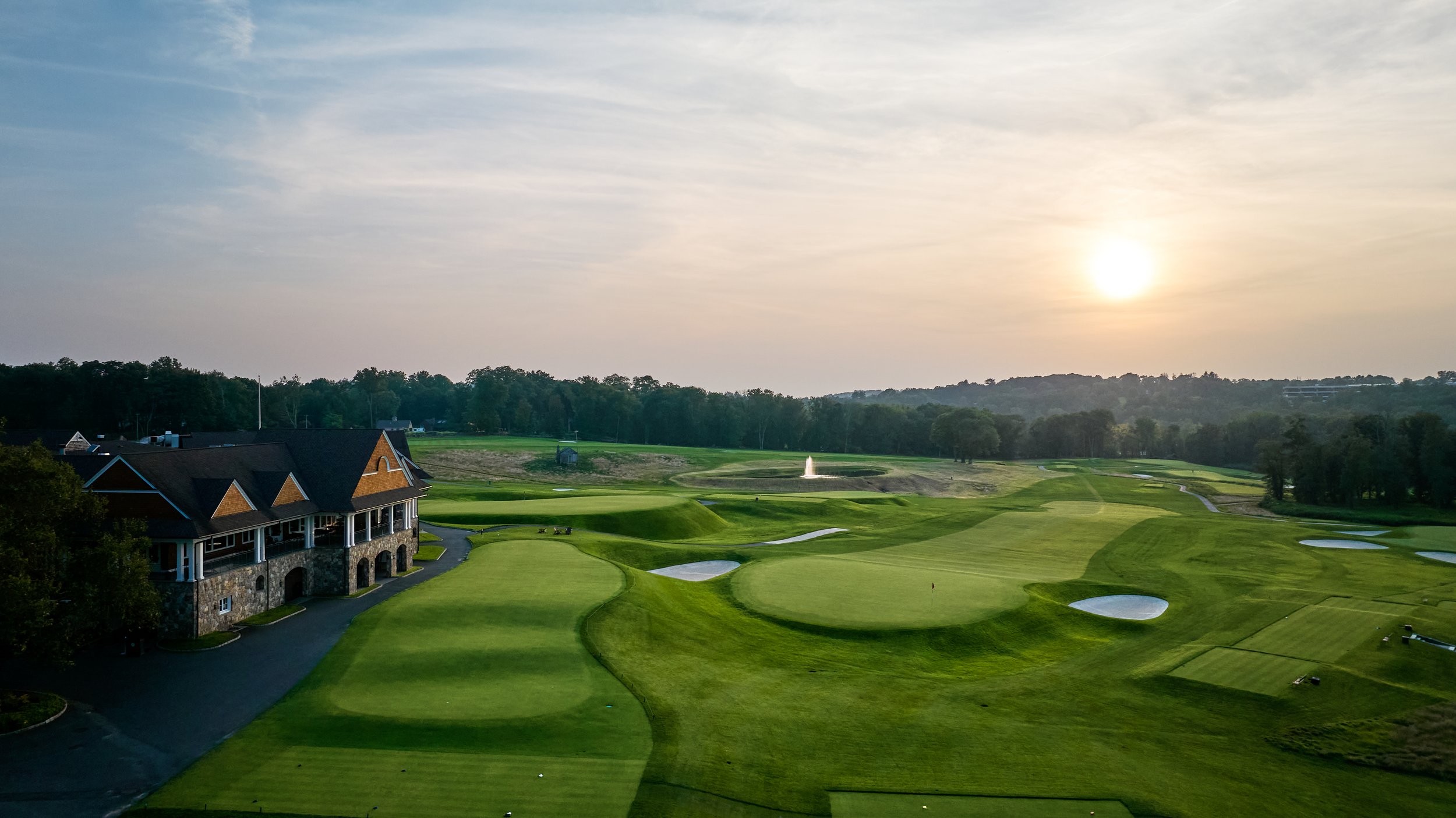 A scenic view of a golf course at sunset, featuring green fairways, sand bunkers, a pond with a fountain, and a clubhouse with a dark roof, trees, and a hill in the background.