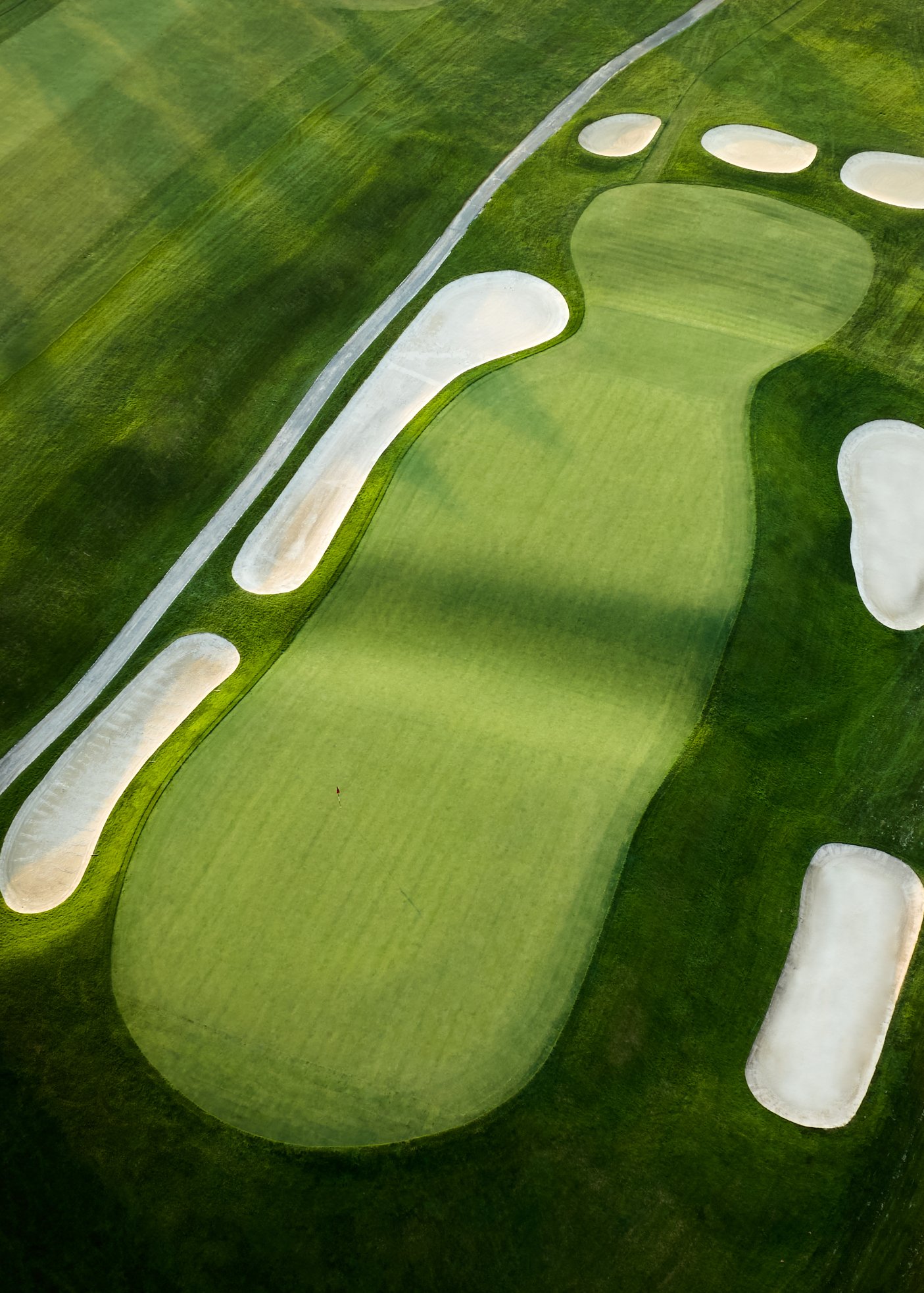 Aerial view of a golf course green with sand bunkers and a flagstick, surrounded by well-maintained grass and a paved pathway.