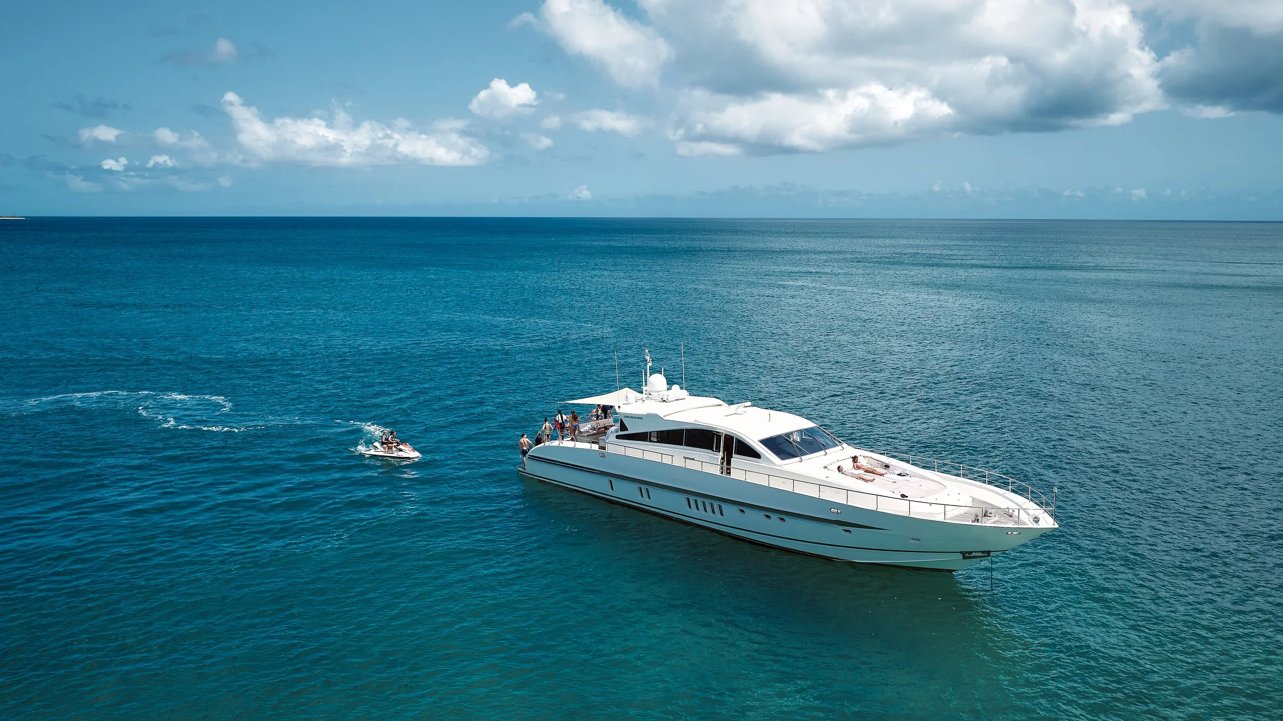 A white yacht on the ocean with a few people on deck and a small jet ski nearby, under a partly cloudy sky.