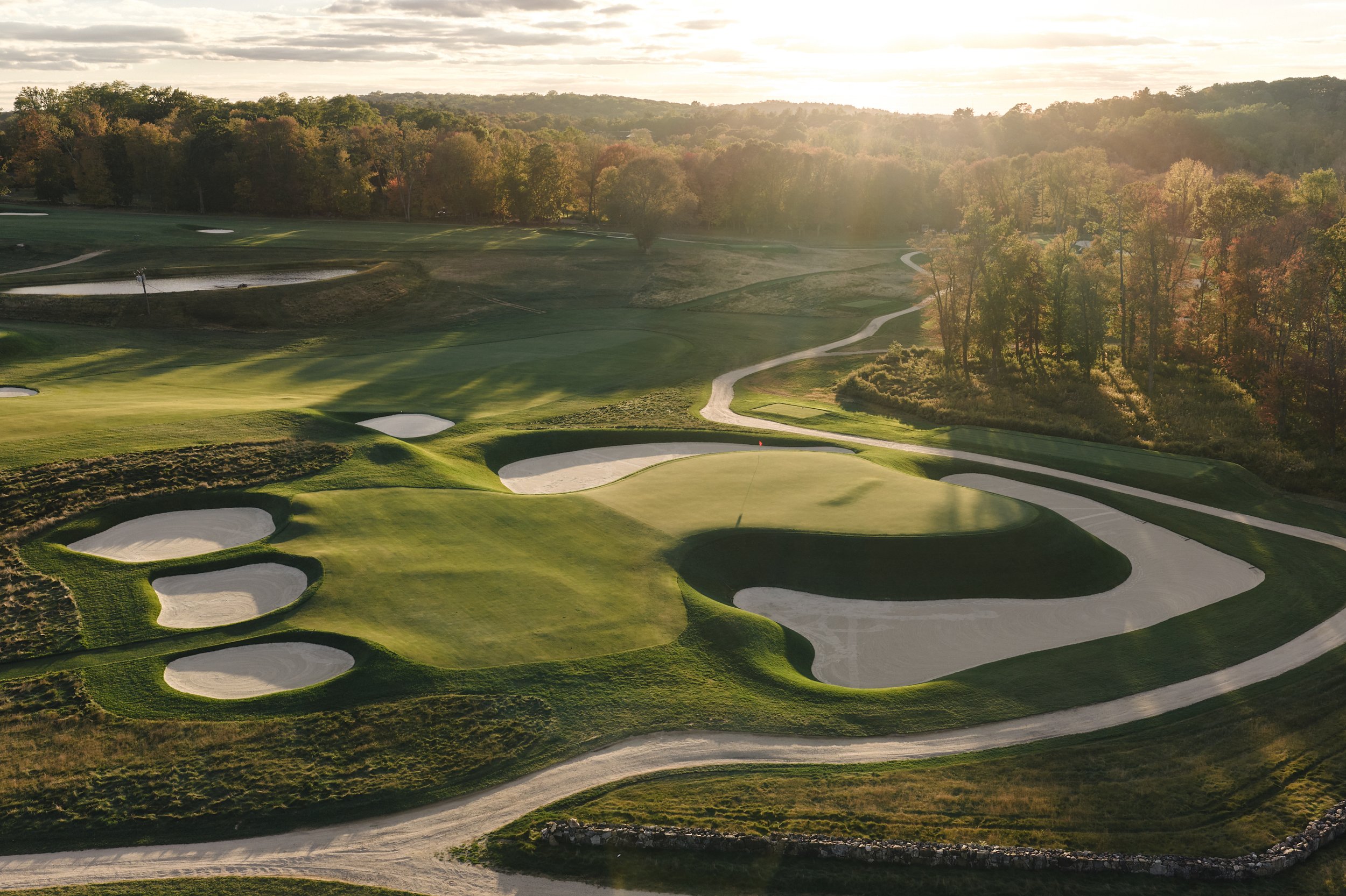 Aerial view of a golf course during sunset with sand traps, green fairways, and trees in the background.