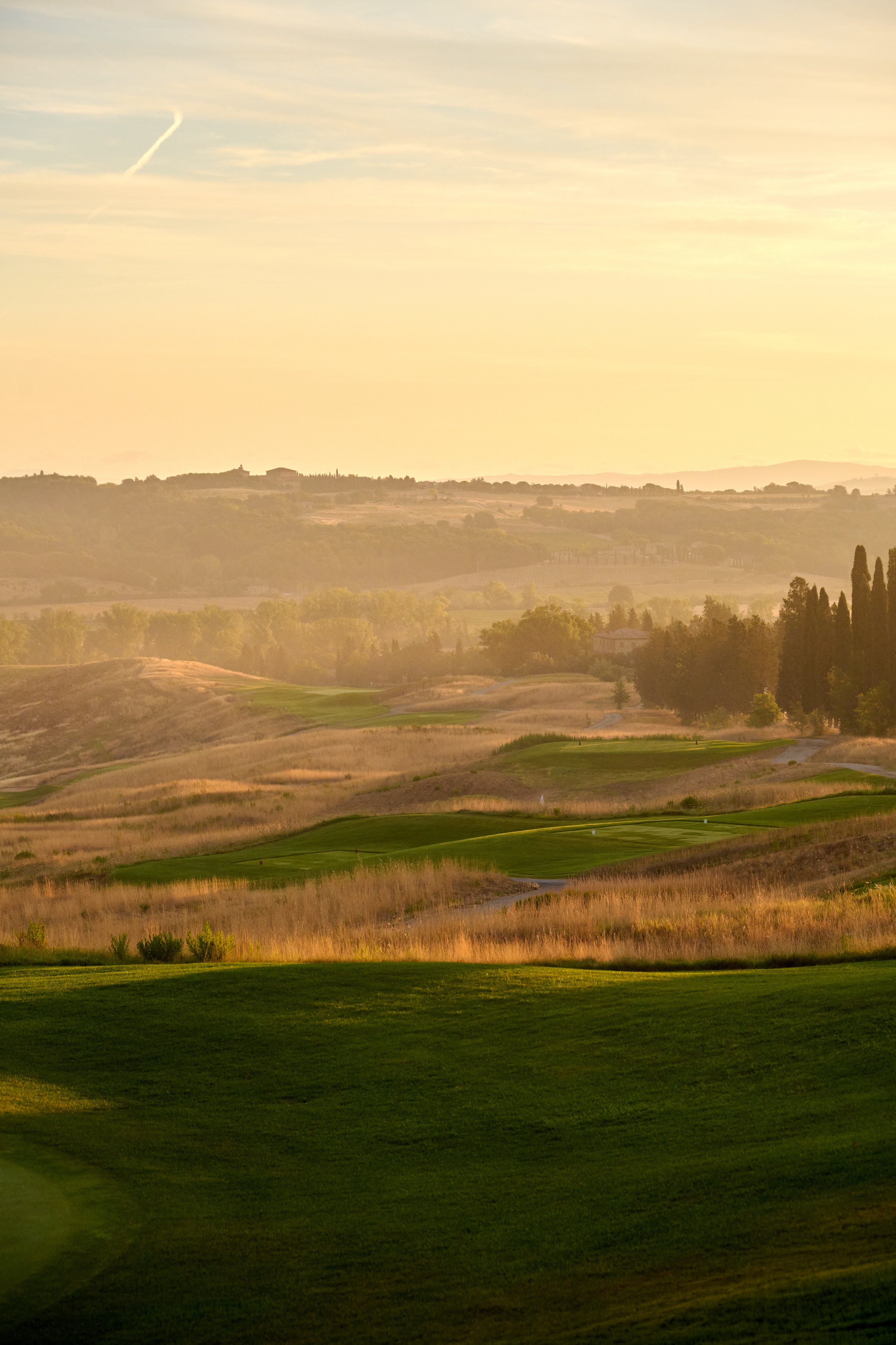 A scenic view of a golf course with green grass, rolling hills, and trees in the distance under a yellowish sky at sunset.