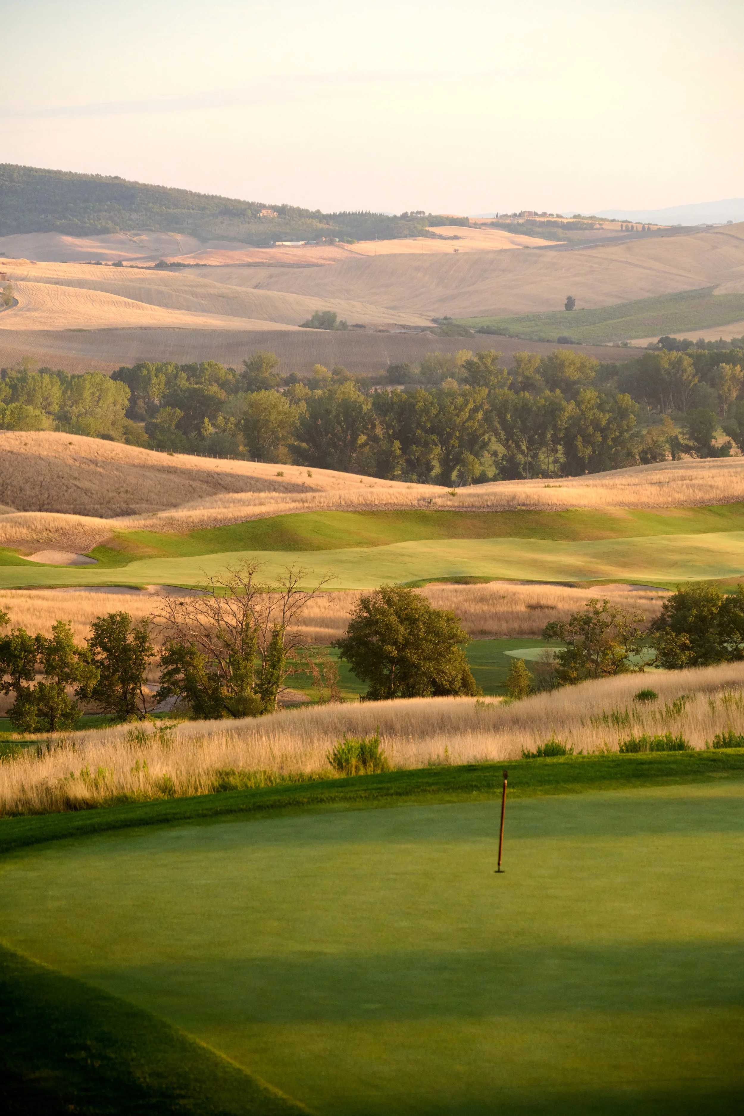 A golf course green with a flag, surrounded by rolling hills and trees in the distance.