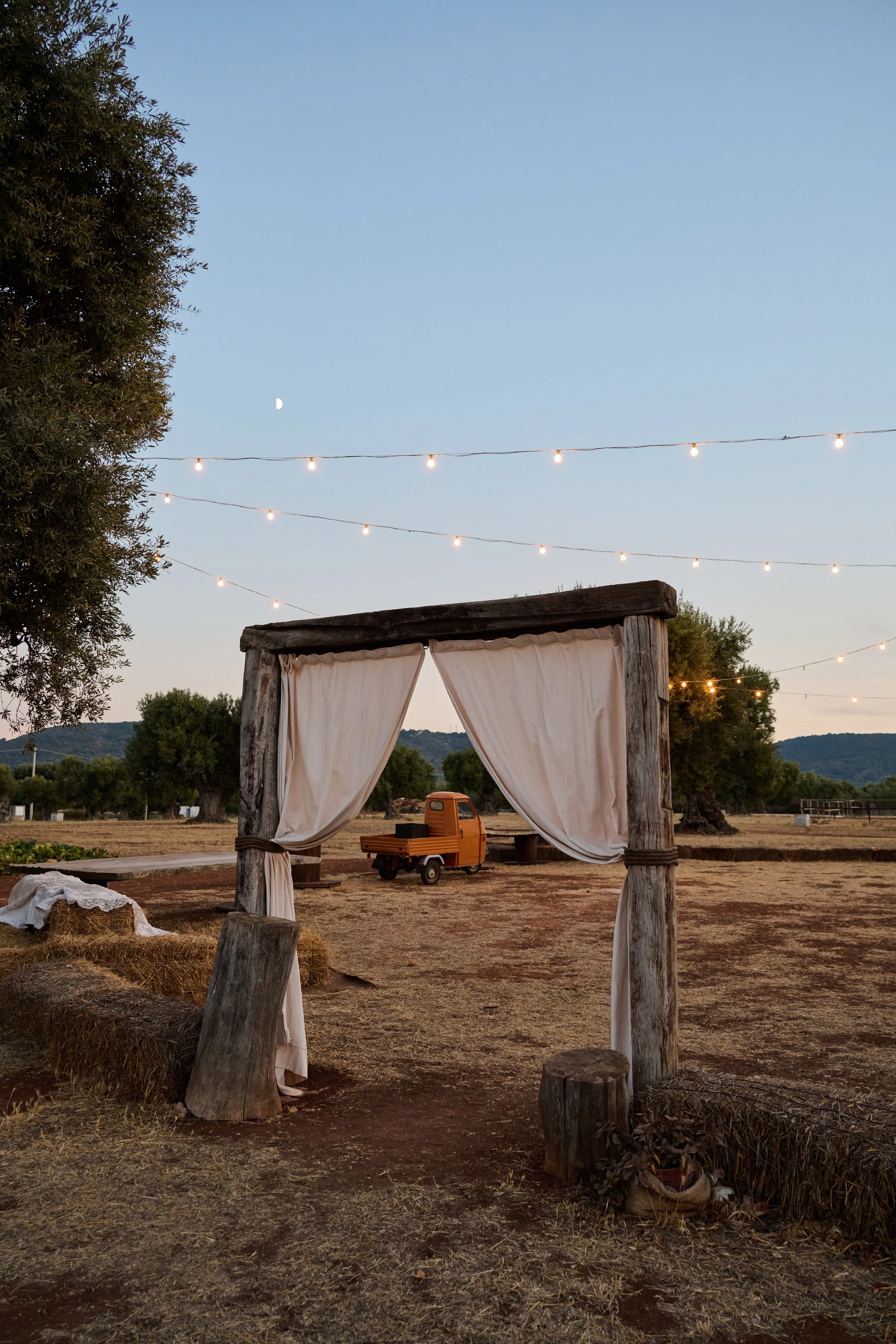 An outdoor rustic wooden archway with white curtains, set in a rural setting at dusk with string lights overhead, a small orange vintage truck in the background, hay bales on either side, and a partly visible large tree on the left.