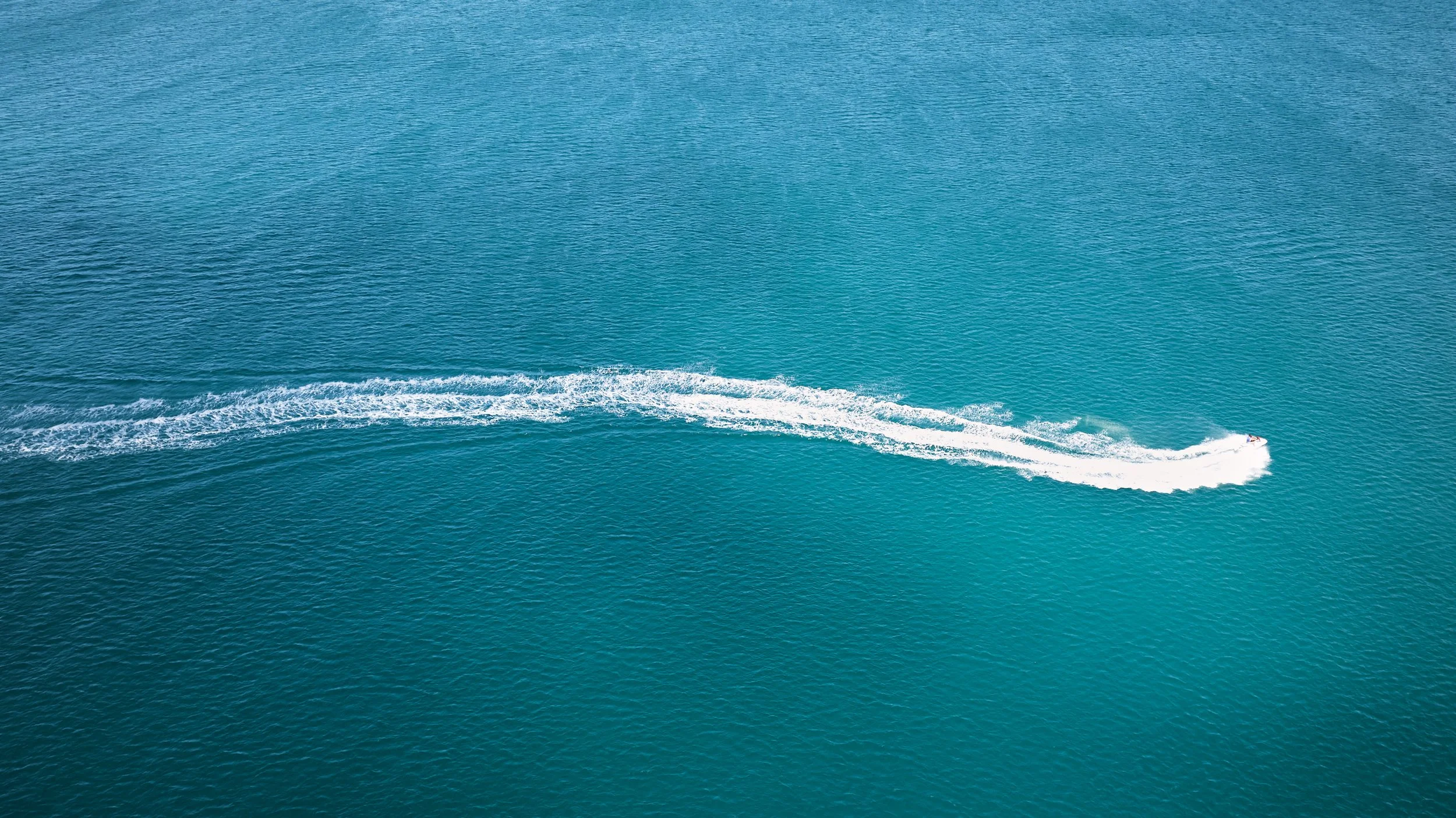 An aerial view of a boat speeding across the water, creating a white wake behind it in a large body of water.