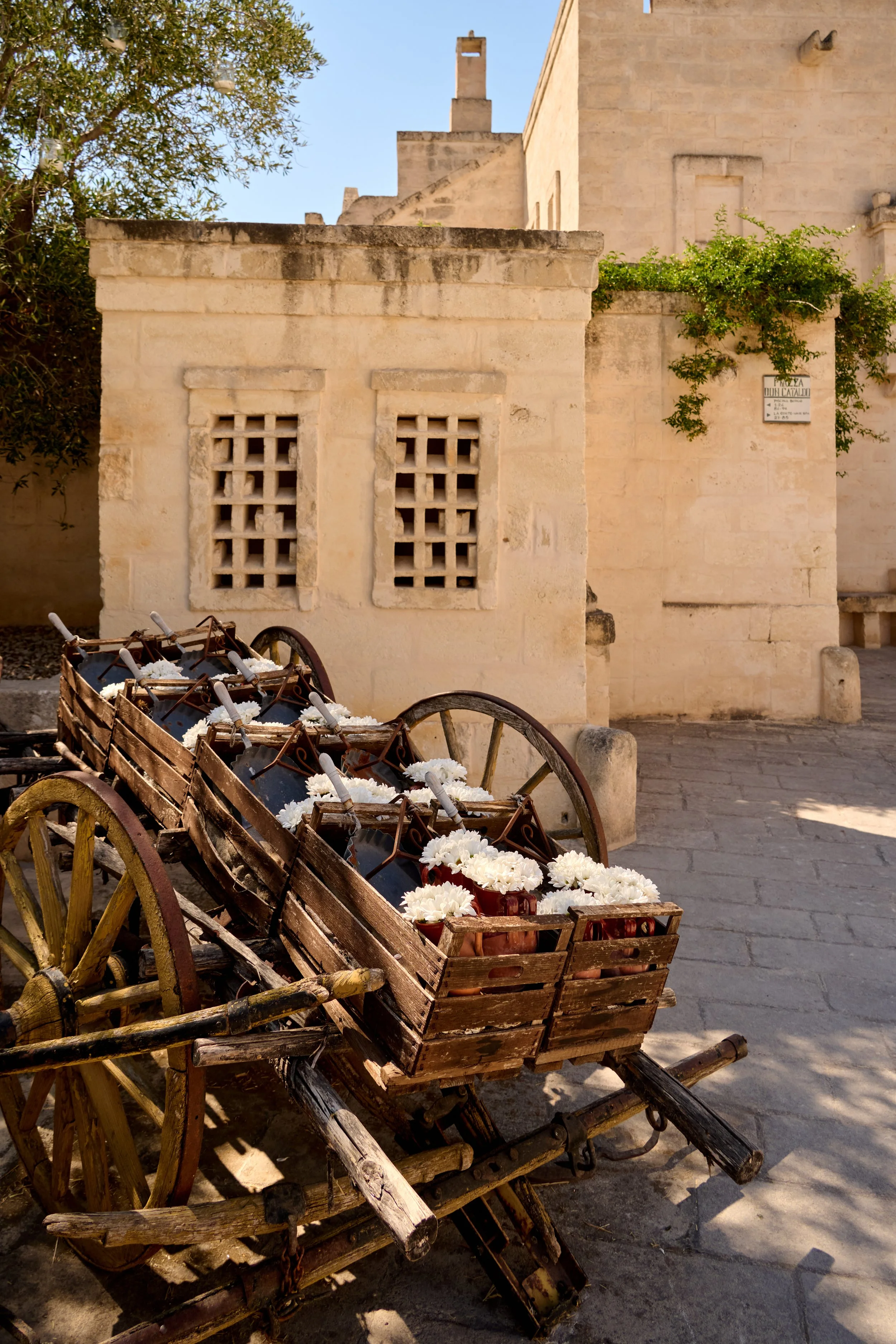 An old wooden cart filled with white flowers parked on a cobblestone street in front of an old stone building with small decorative windows and a sign. Greenery is growing on the building wall.