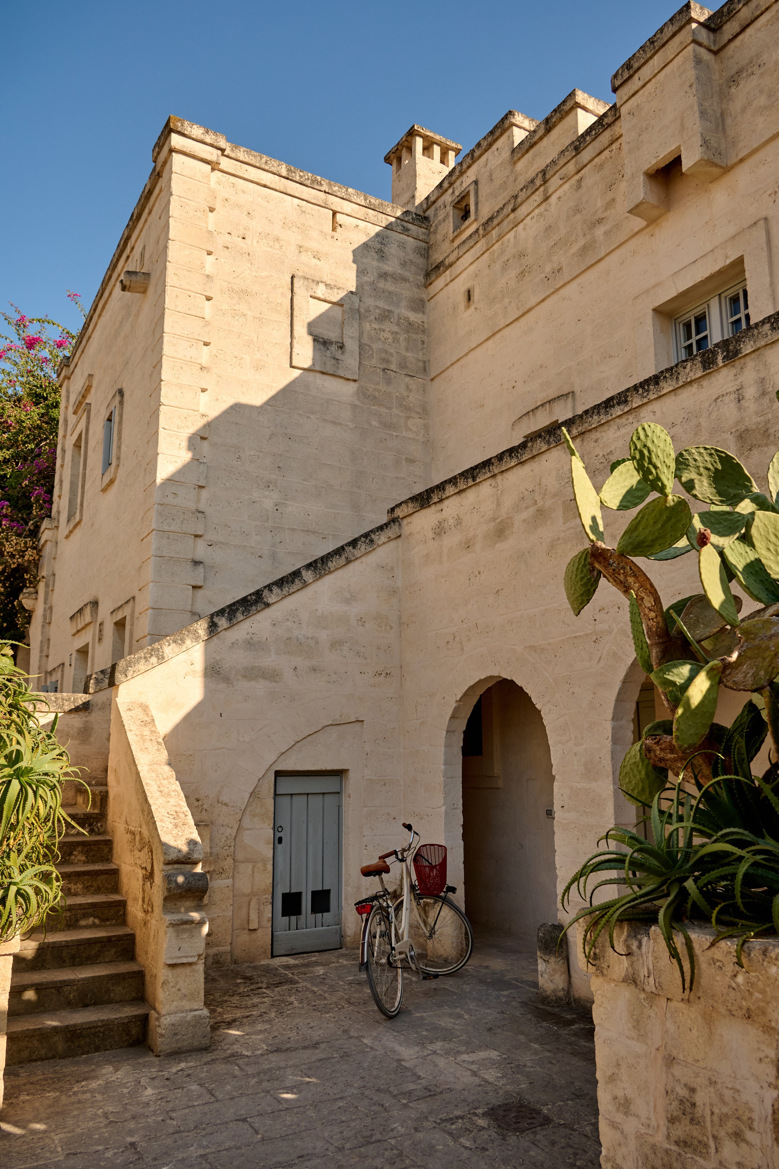 A limestone building with stairs, an arched entryway, a bicycle, and cacti in an outdoor courtyard under a clear sky.