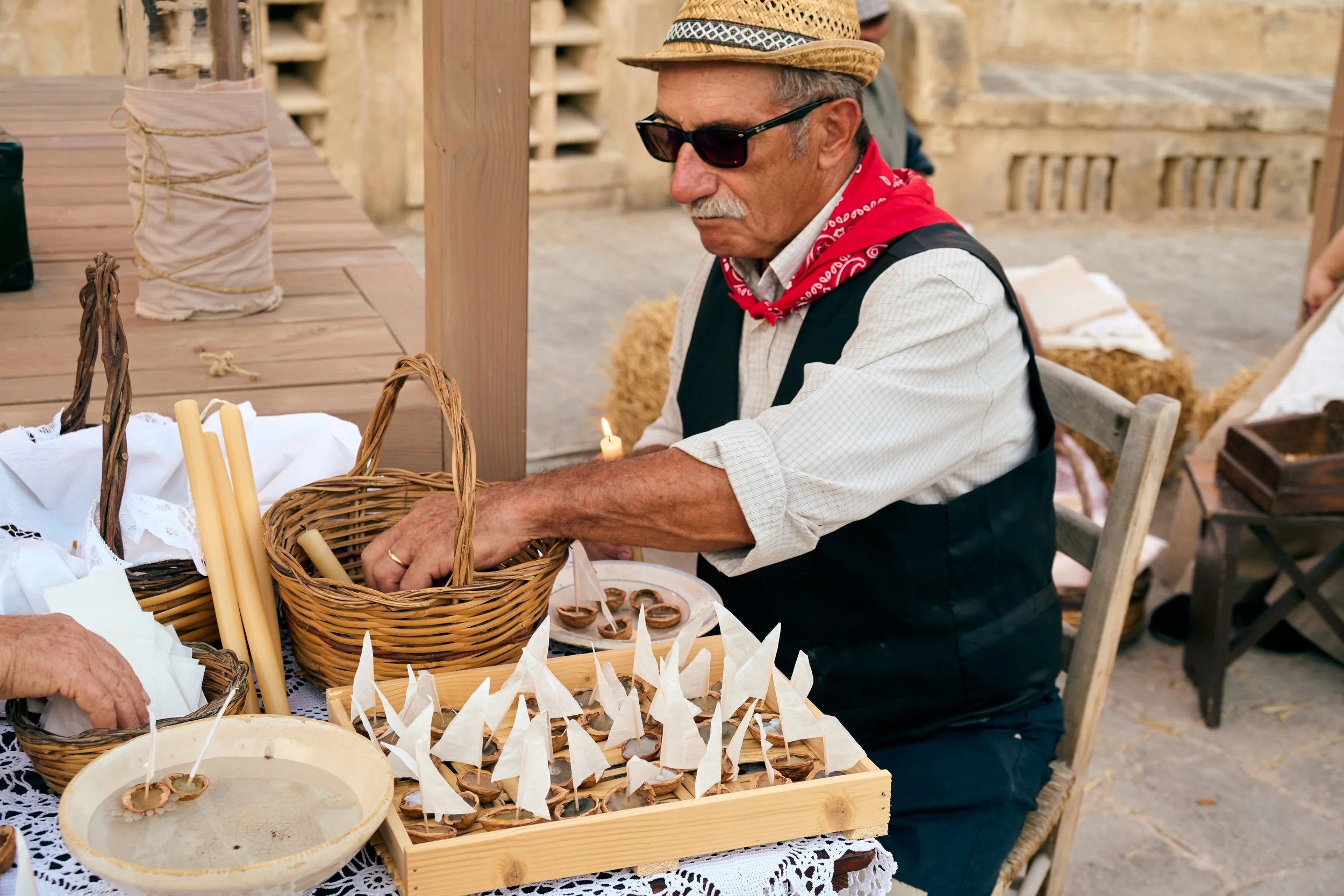 An older man wearing sunglasses, a straw hat, a red bandana, a light shirt, and a dark vest is sitting at a table with woven baskets and trays of small dishes covered with paper flags. The setting appears to be outdoors, possibly at a market or event