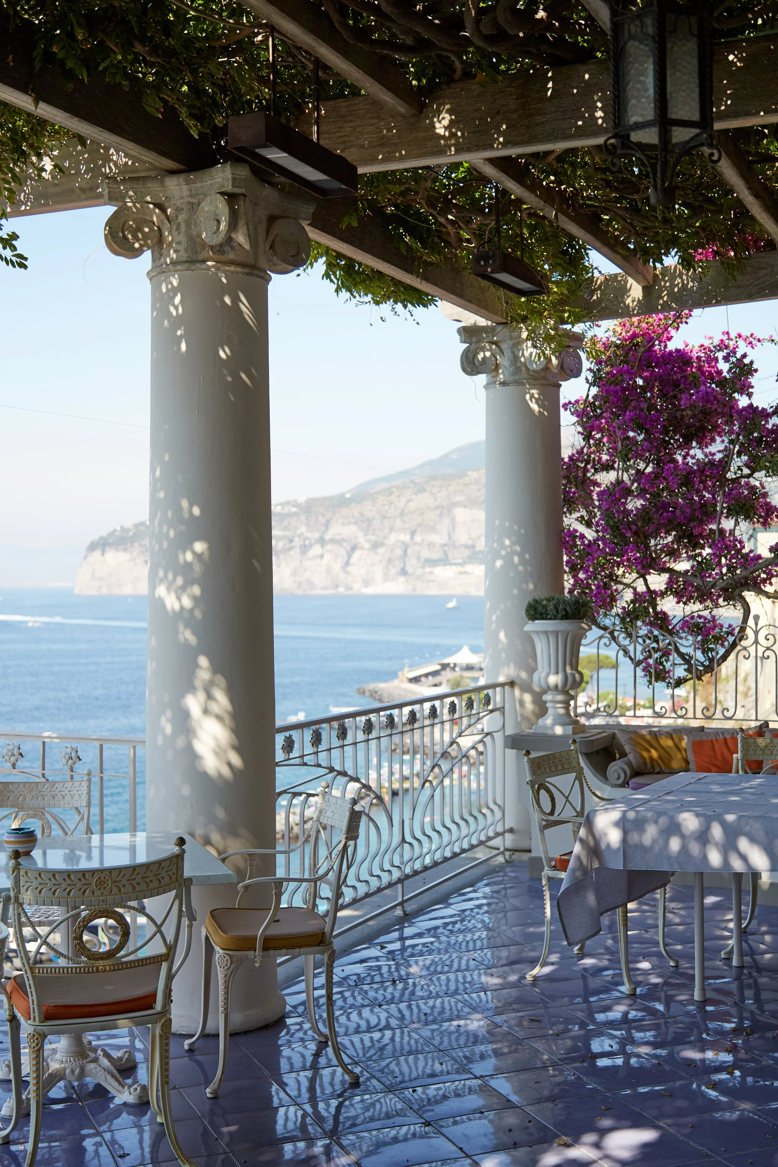 A seaside terrace with white ornate chairs and tables, shaded by a pergola with lush foliage, overlooking the ocean and cliffs in the distance.