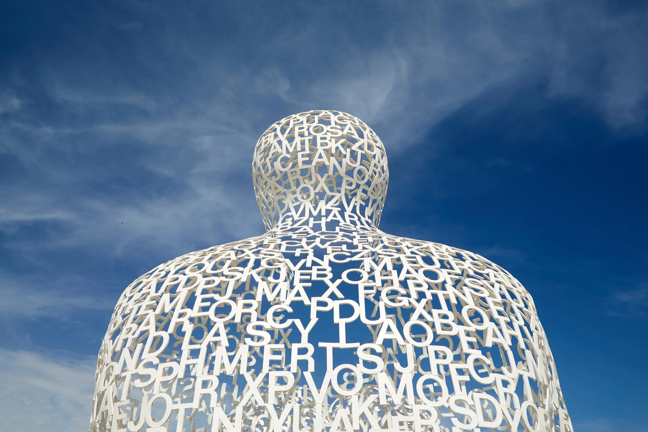 Large sculpture of a human figure made of interconnected white letters, against a blue sky with some clouds.