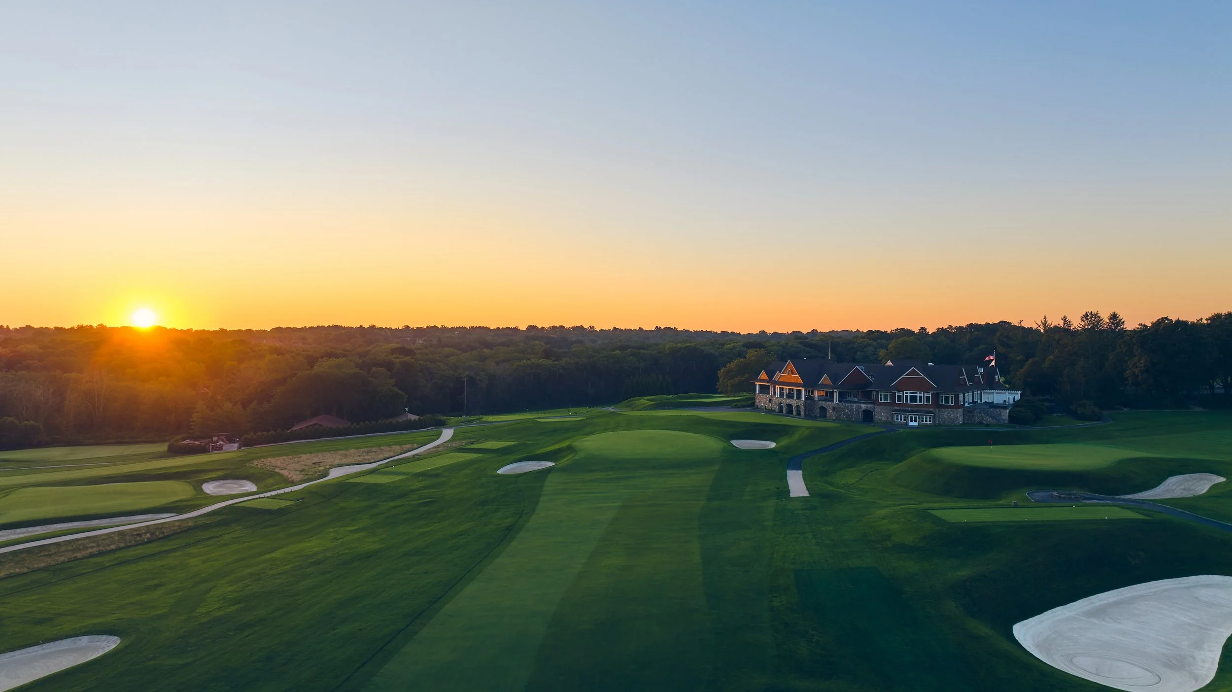 Sunset over a golf course with green fairways, sand traps, and a large clubhouse in the background.