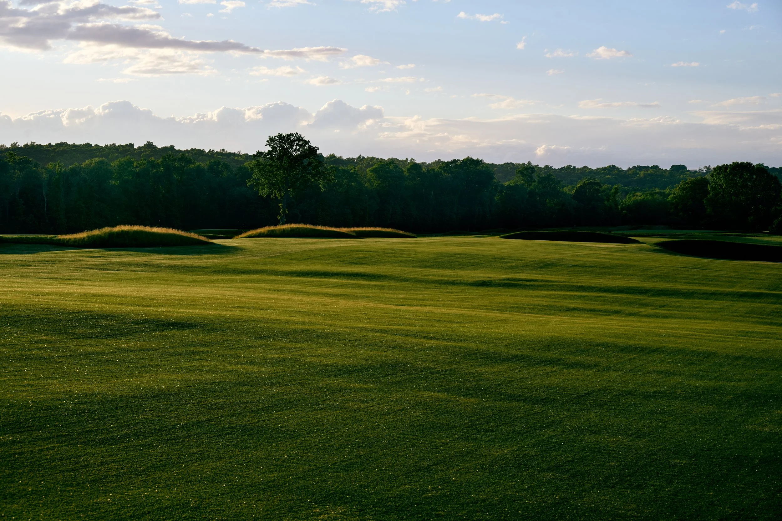 A golf course with neatly mowed grass, sand traps, and a backdrop of trees under a partly cloudy sky.