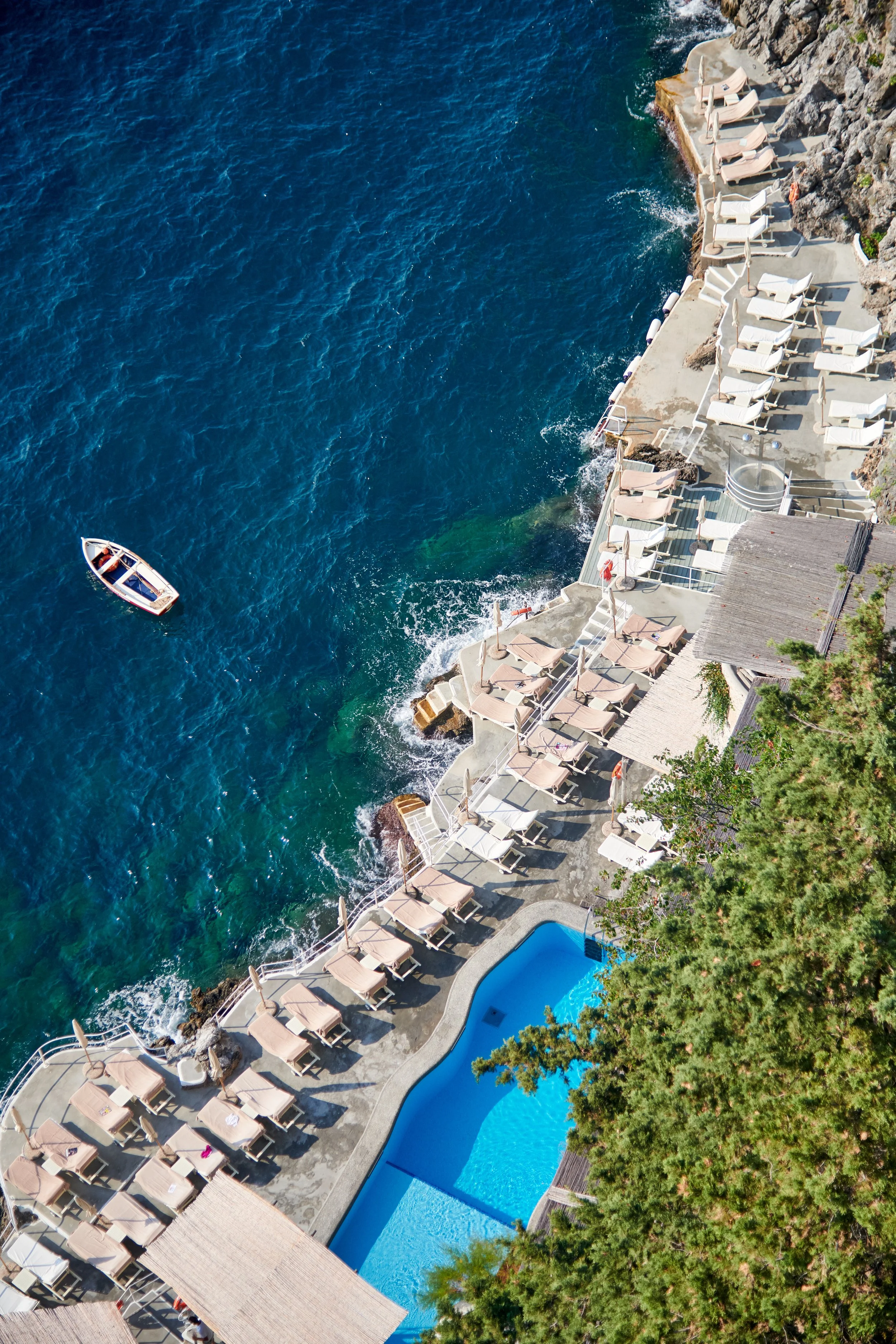 Aerial view of a seaside resort with a swimming pool, lounge chairs, and umbrellas along the rocky shoreline, with a boat in the water.