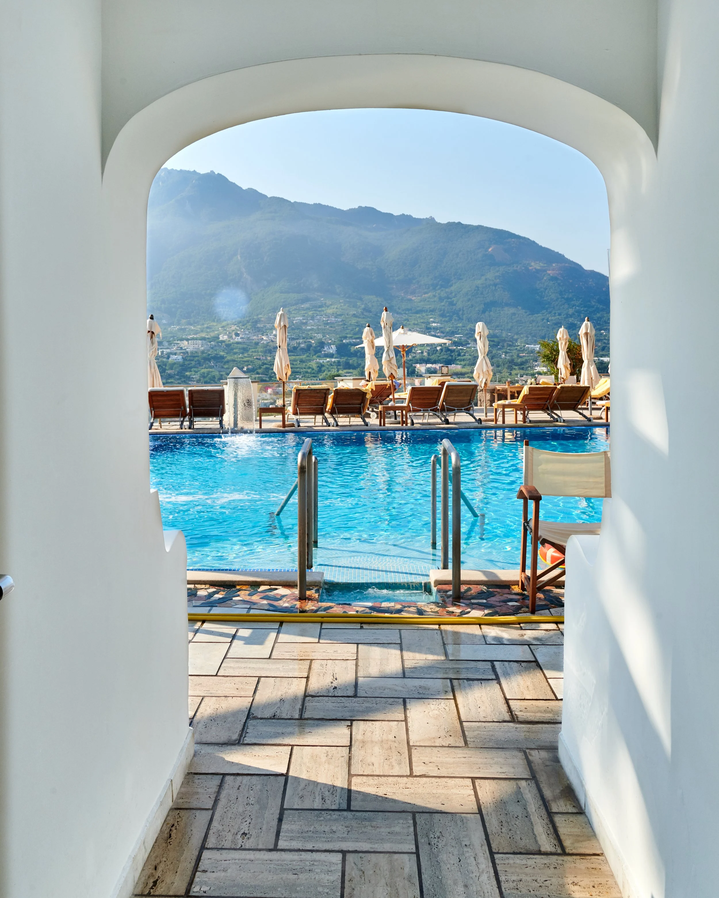 View of a swimming pool with lounge chairs and umbrellas, framed by a white archway, mountain landscape in the background.