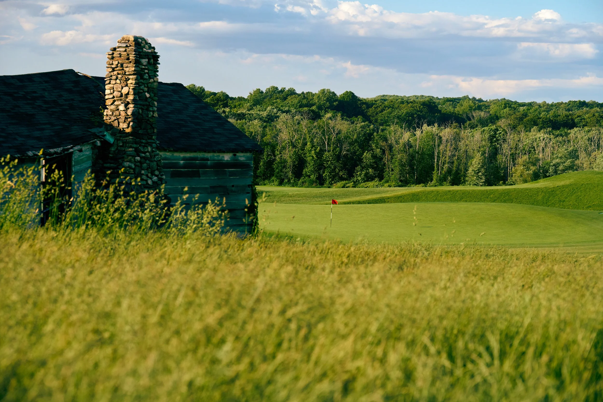 A golf course with a green and a red flag, a small wooden building with a stone chimney, surrounded by lush green grass and trees in the background under a partly cloudy sky.